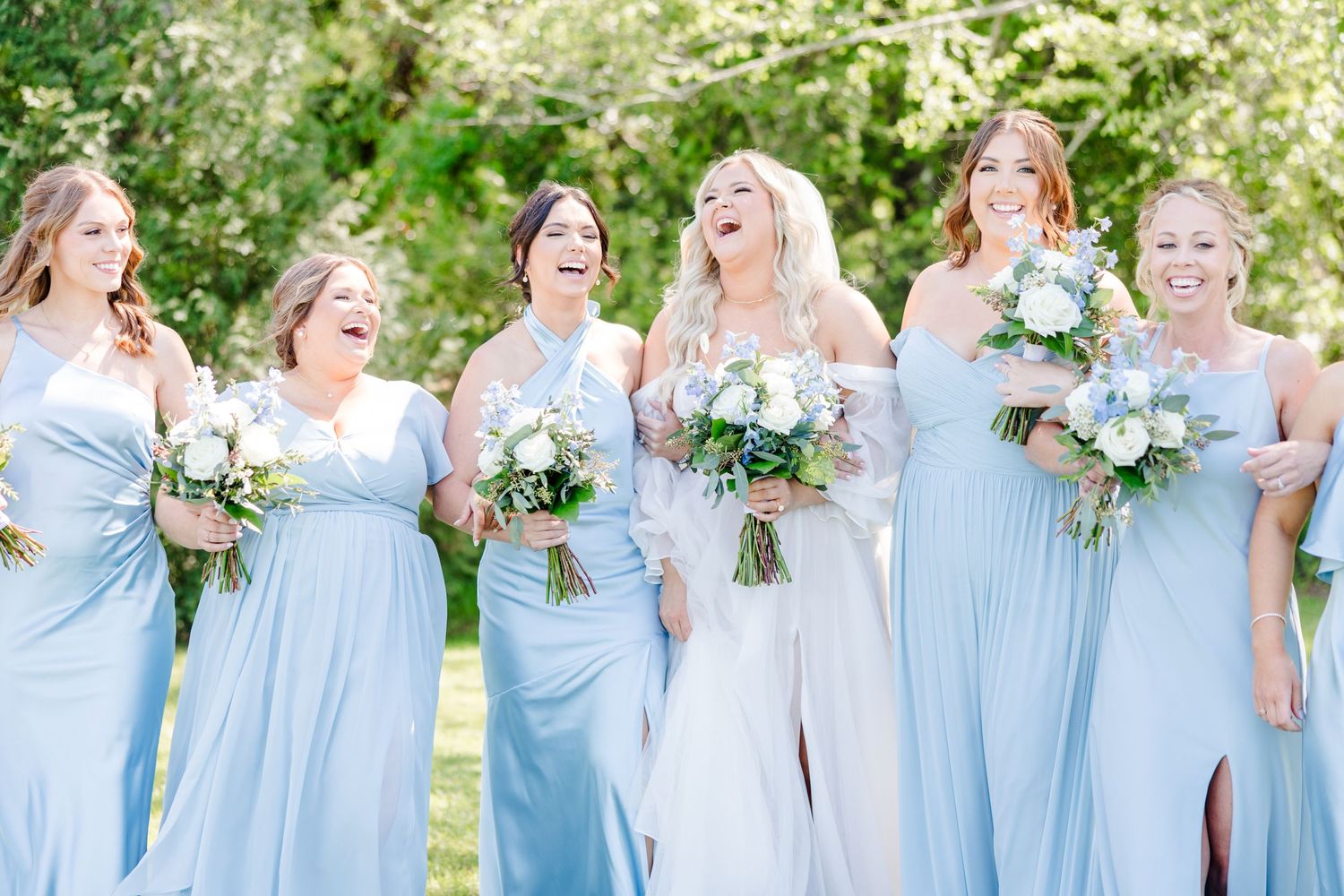 Bridesmaids in flowing light blue dresses hold white and green bouquets while laughing together outdoors.
