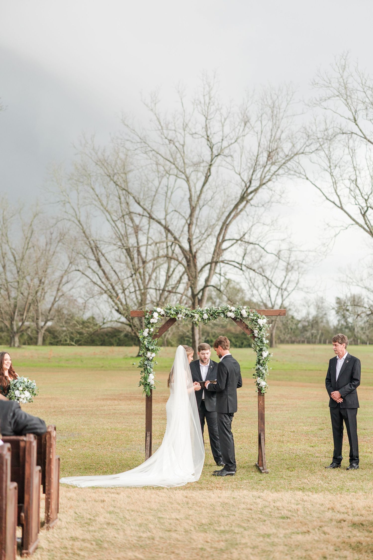 Outdoor wedding ceremony under floral arch in open field with bride, groom, and officiant on overcast day.
