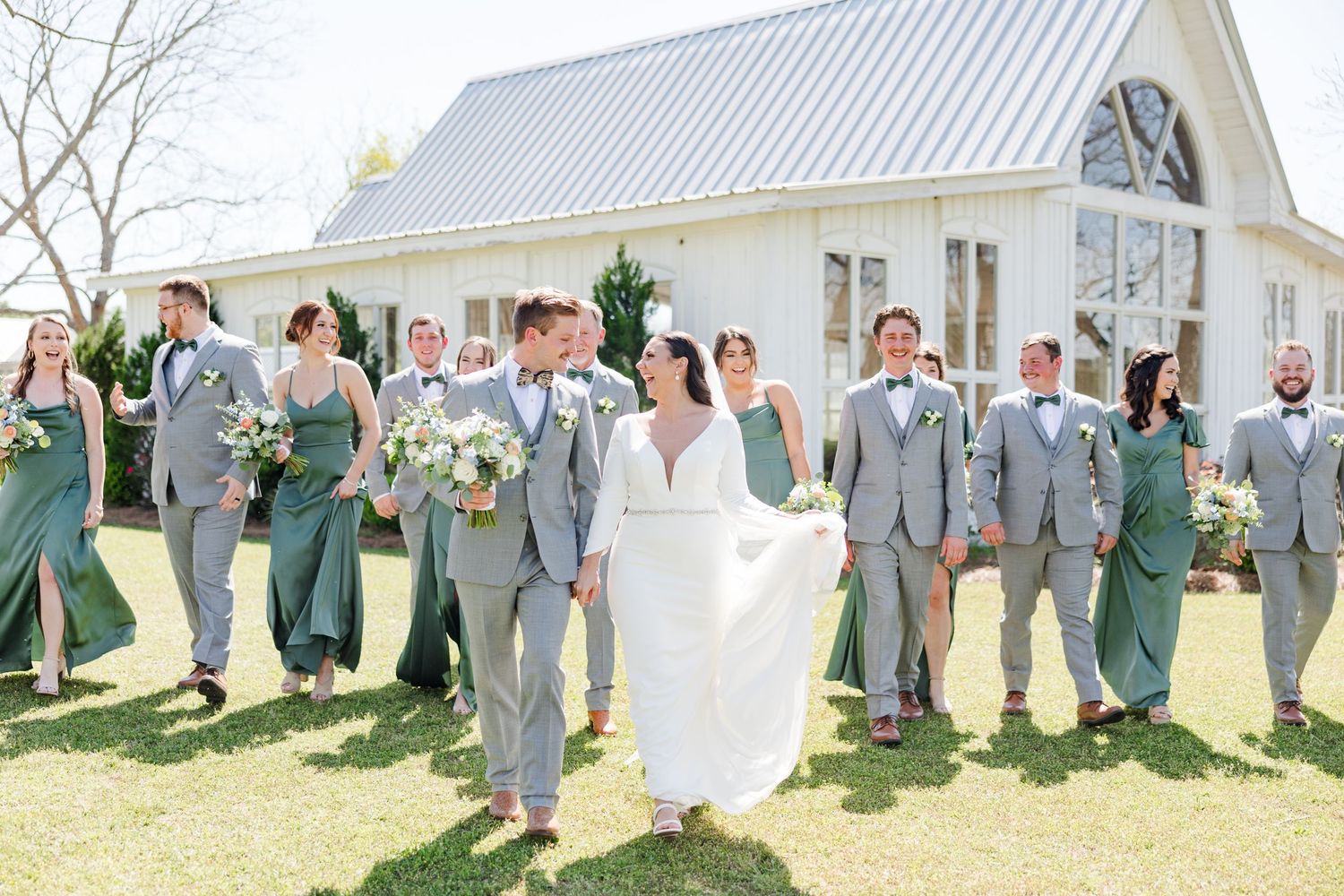 Large wedding party poses in front of white barn with groomsmen in gray and bridesmaids in green.
