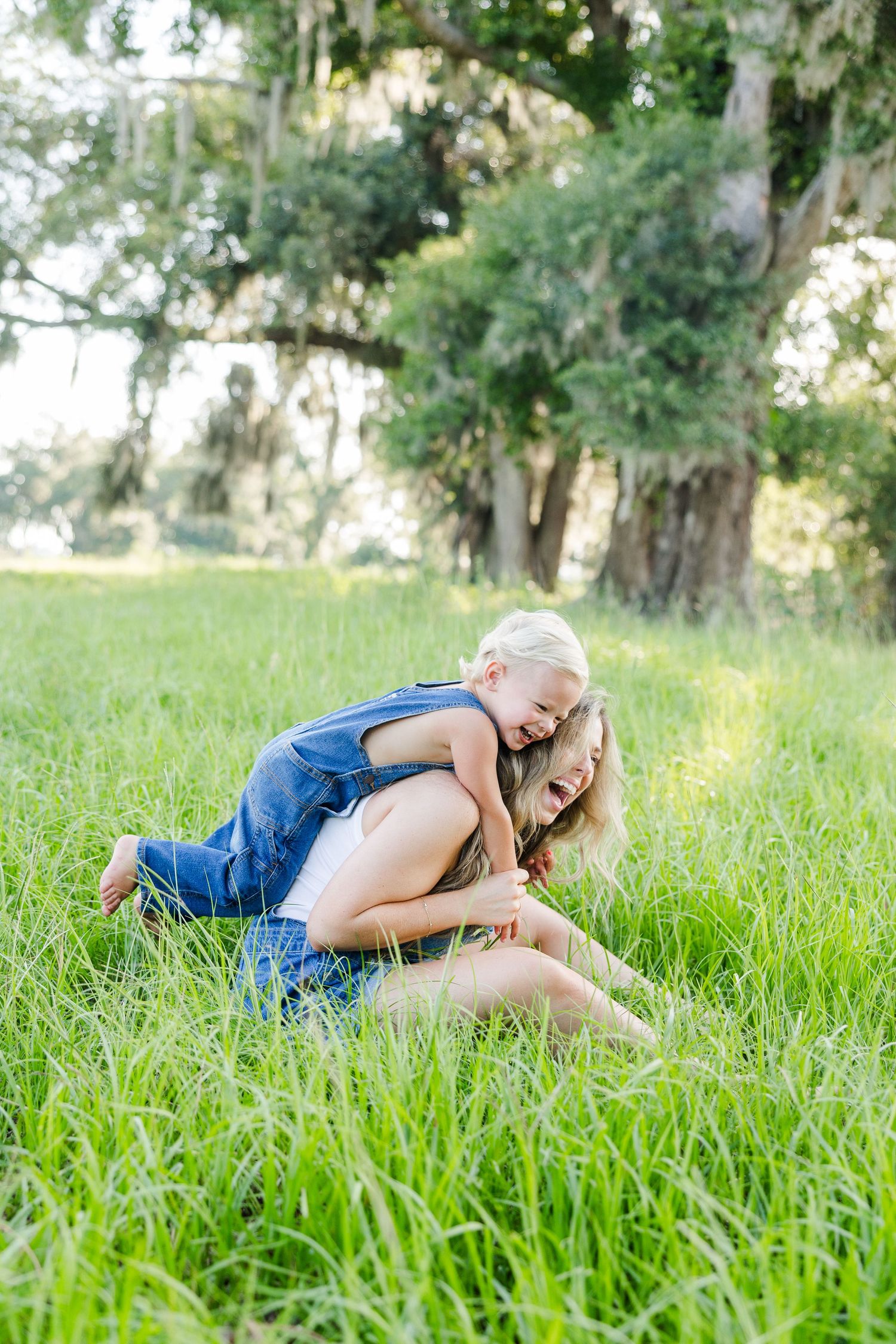 A mother and young child playfully tumbling together in lush green grass beneath moss-covered oak trees.