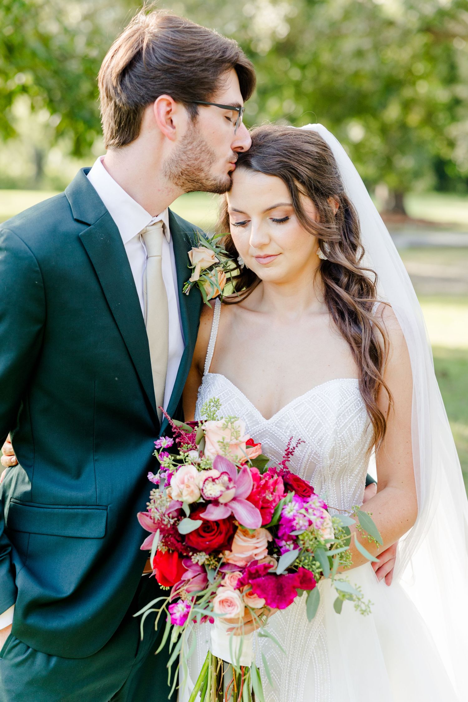Groom in green suit kisses bride on forehead as she holds vibrant pink and red wedding bouquet outdoors.