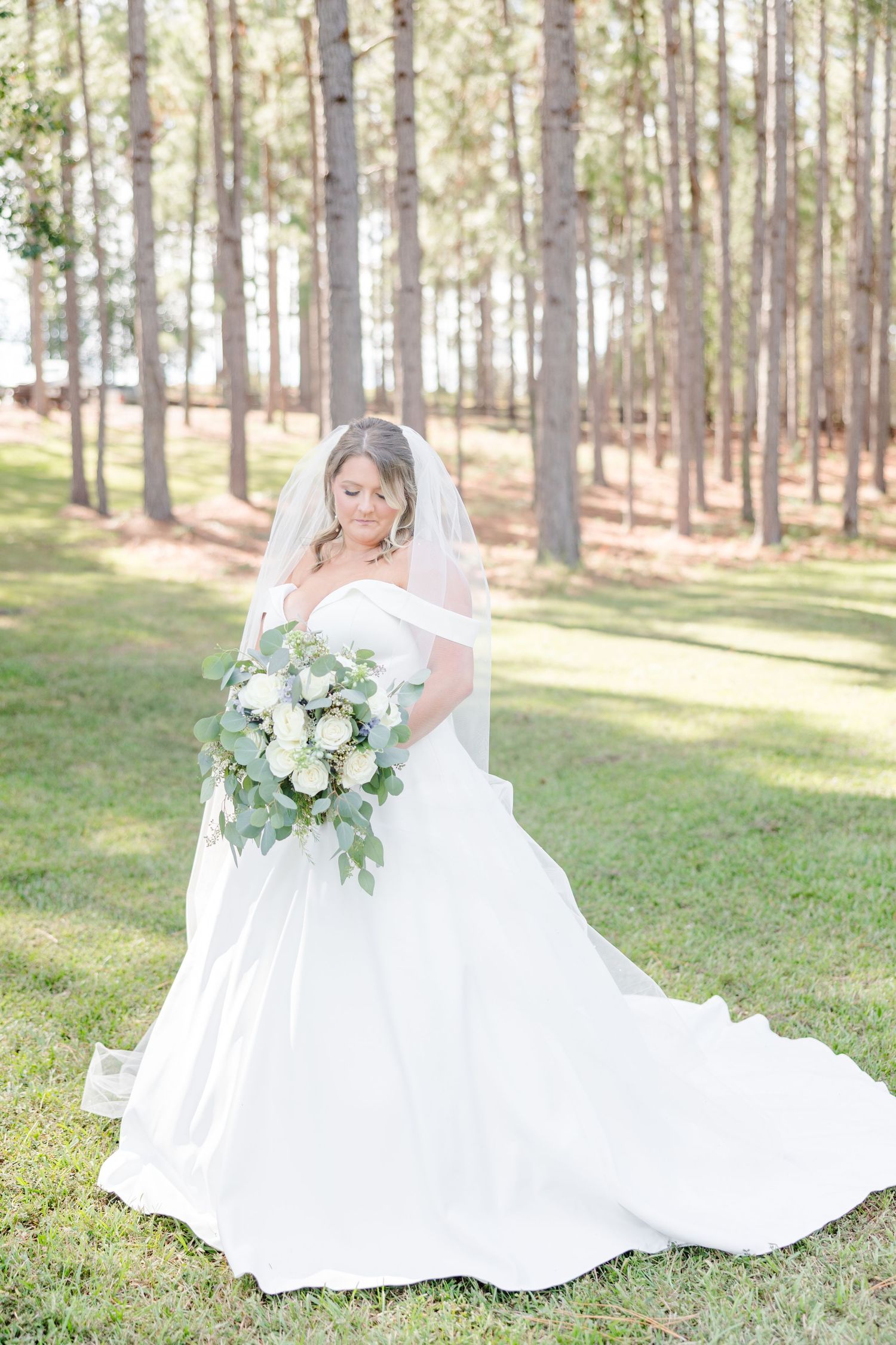 Elegant bridal portrait showcasing flowing white dress and lush greenery bouquet in forest setting.