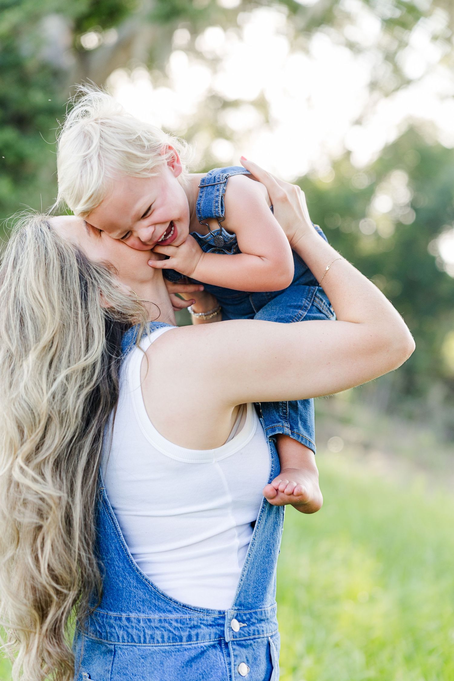 A mother with long blonde hair lifts her laughing toddler outdoors, both wearing denim overalls in a sunny green park.