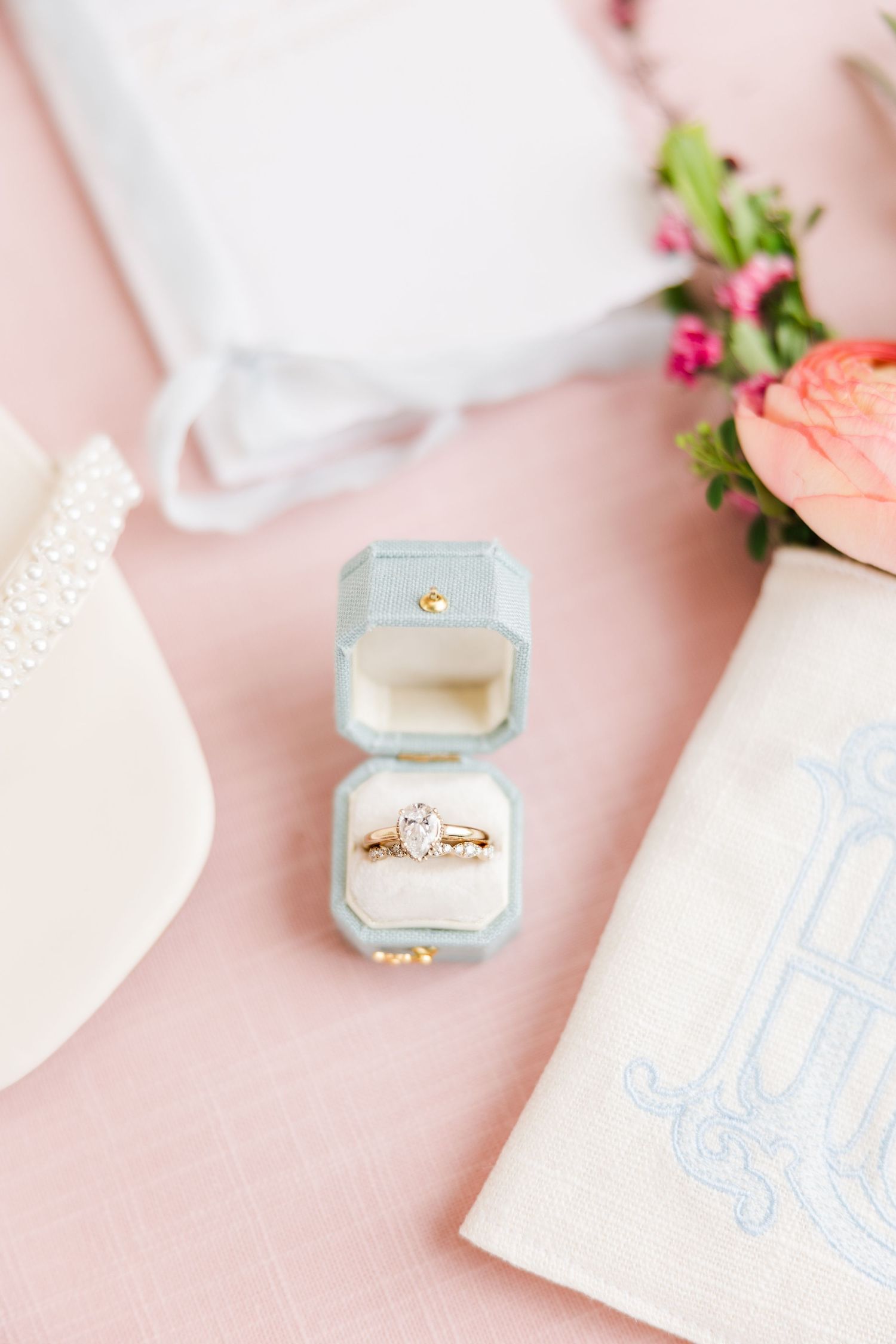 Gold diamond engagement ring and wedding band displayed in a light blue velvet ring box on a soft pink background.