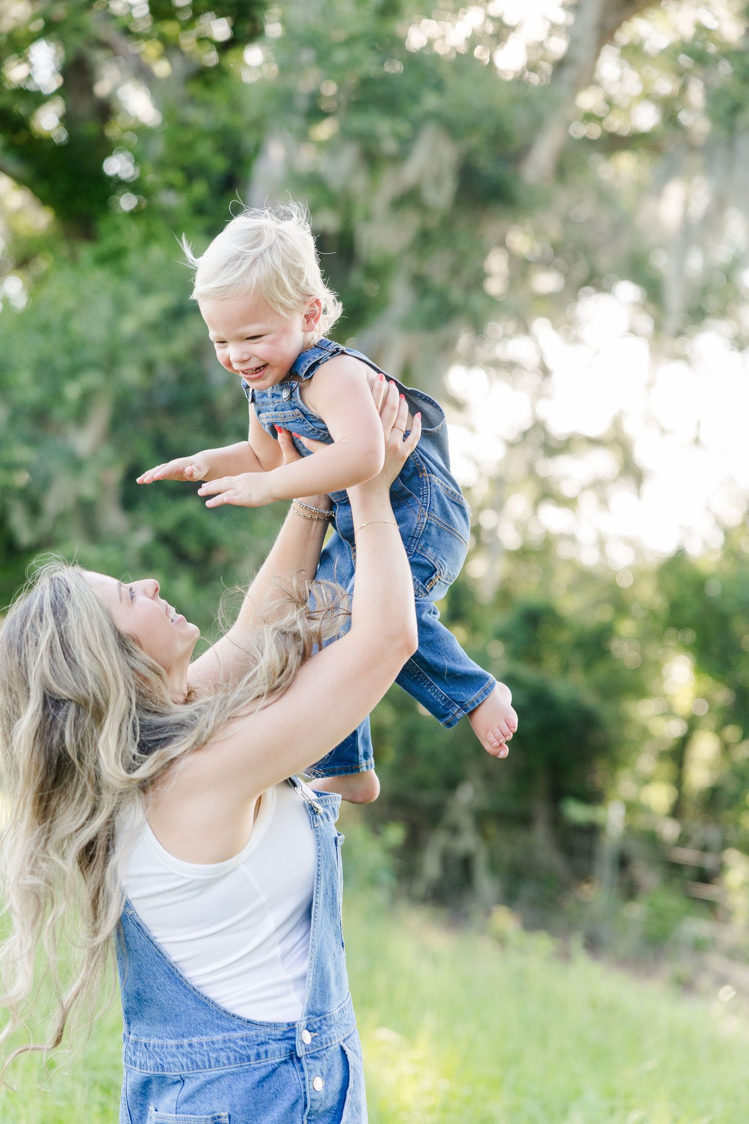 A joyful mother lifts her laughing toddler in the air outdoors, both wearing denim outfits in a lush green setting.