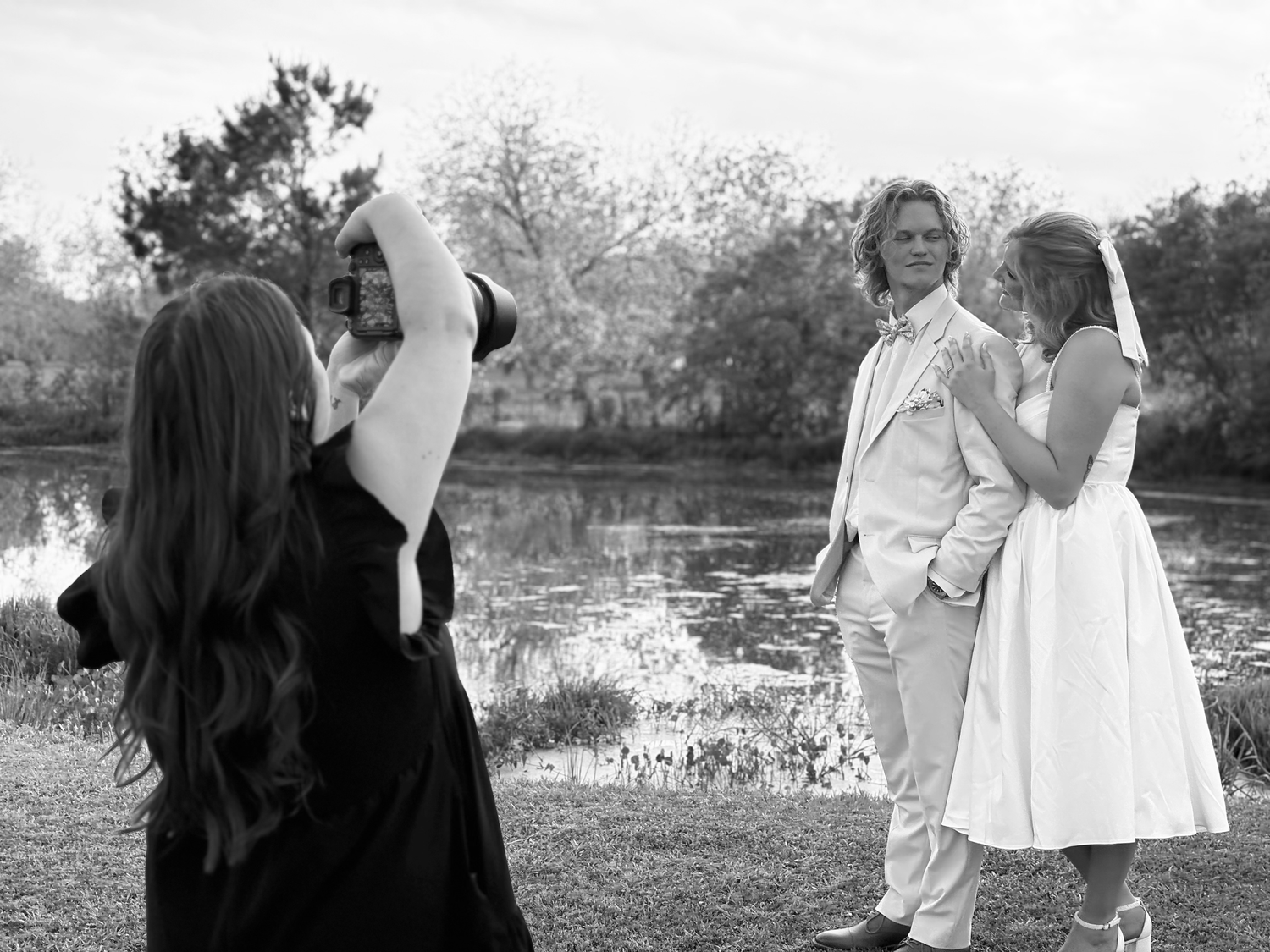 Black and white photo of a photographer capturing a wedding couple outdoors near a pond.