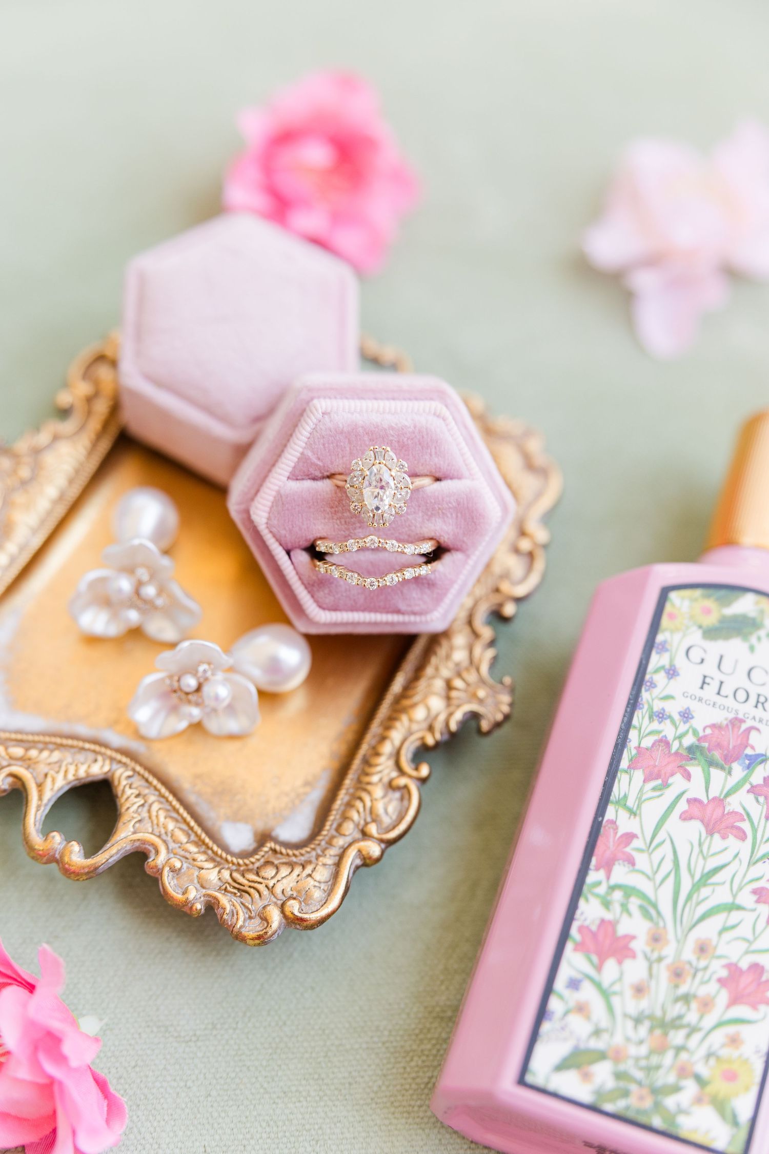 Pink velvet hexagon ring box with diamond engagement ring on gold tray beside Gucci Flora perfume and pearl earrings.