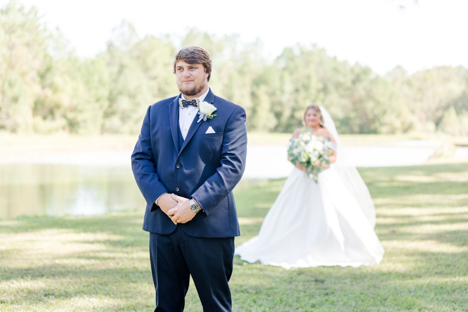 Wedding party members pose on grass with bride in white dress visible in background.