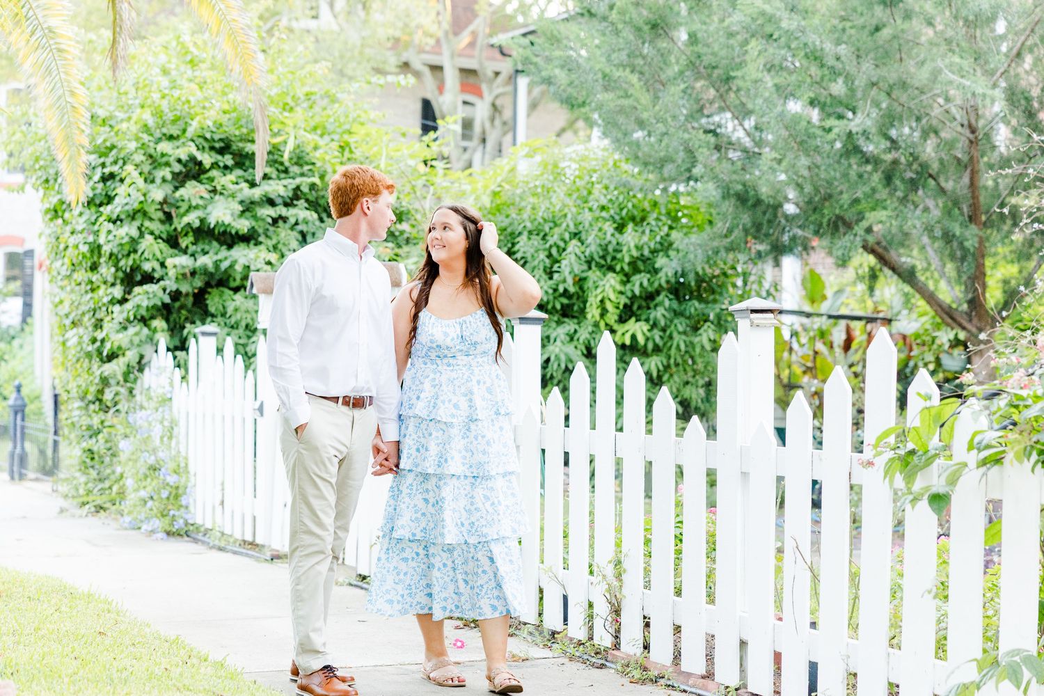 Couple walks along a white picket fence path, woman in blue floral dress, man in white shirt and khakis, lush greenery.