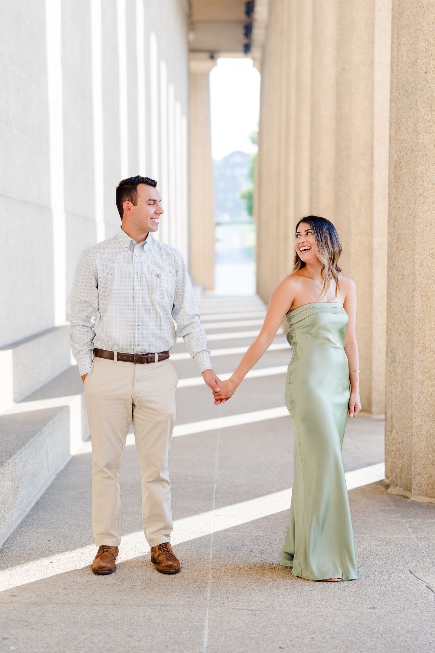 Couple holding hands smiling at each other, woman in sage green gown, man in khakis, standing between stone columns.
