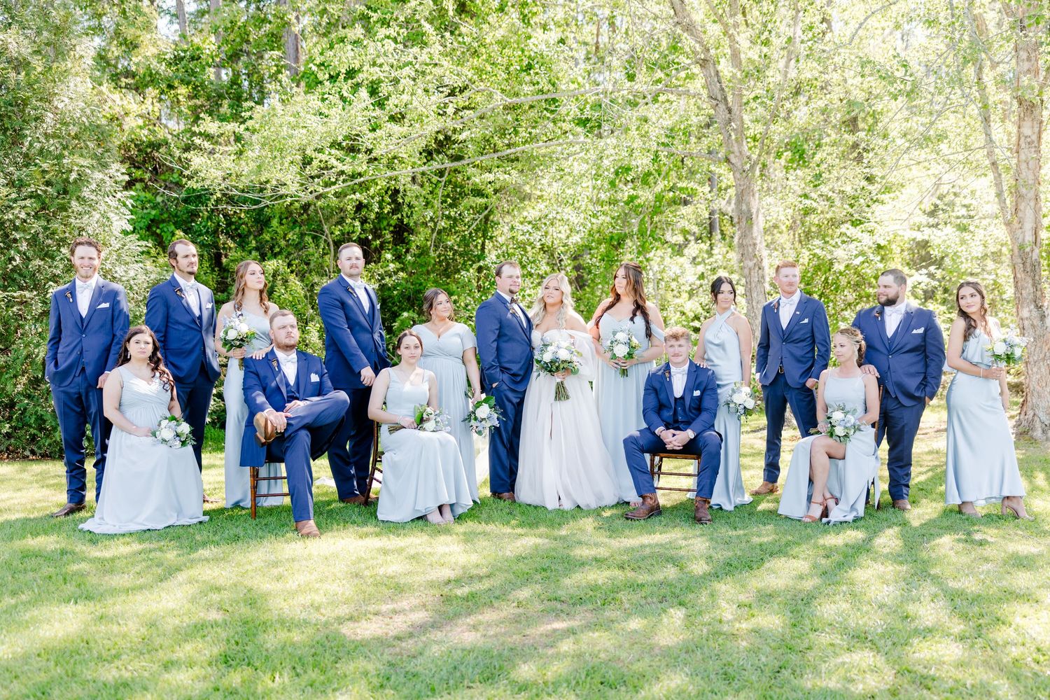 Large wedding party group photo with navy suits and light blue dresses against green foliage.
