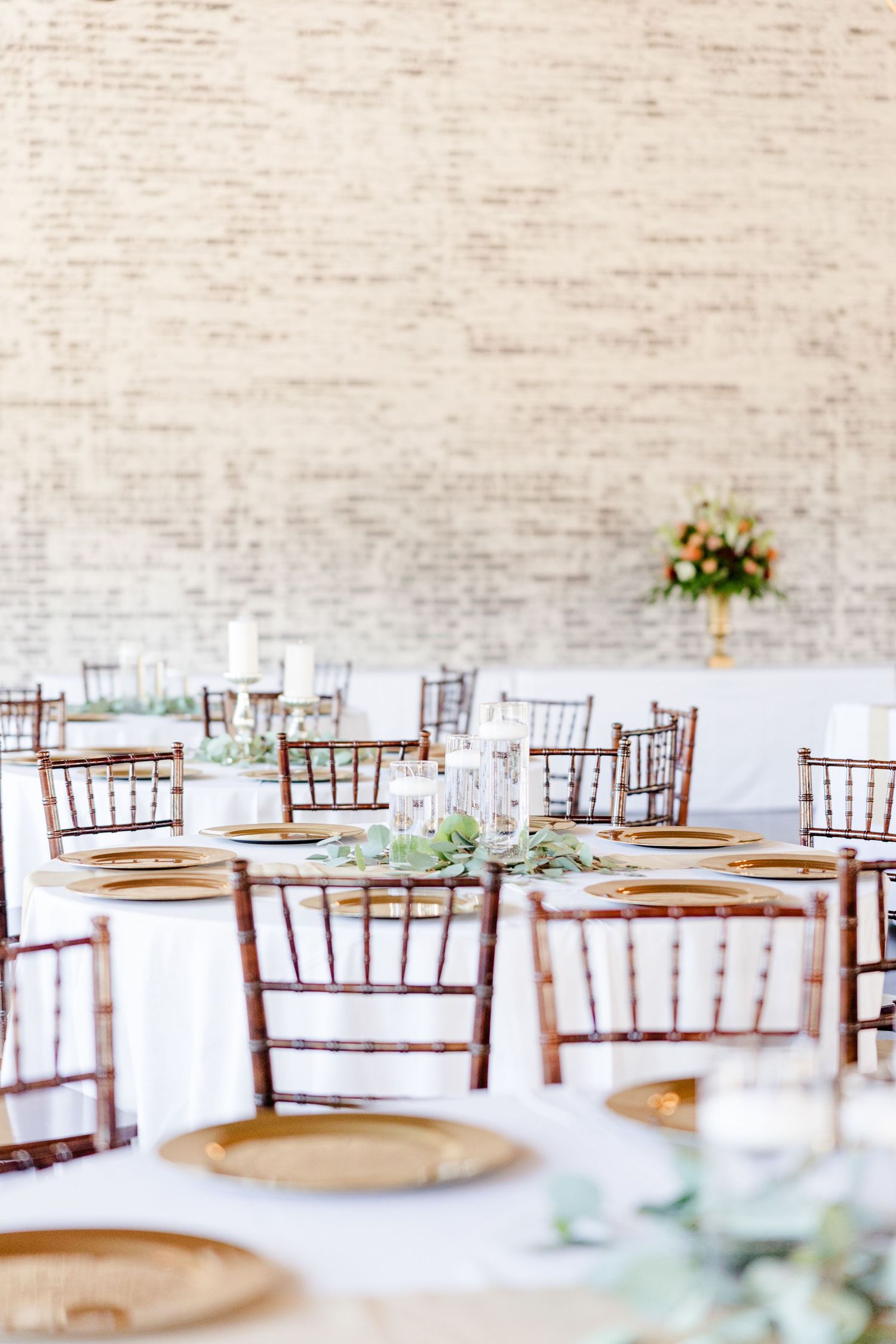 Elegant reception setup with chiavari chairs and place settings against white brick wall.