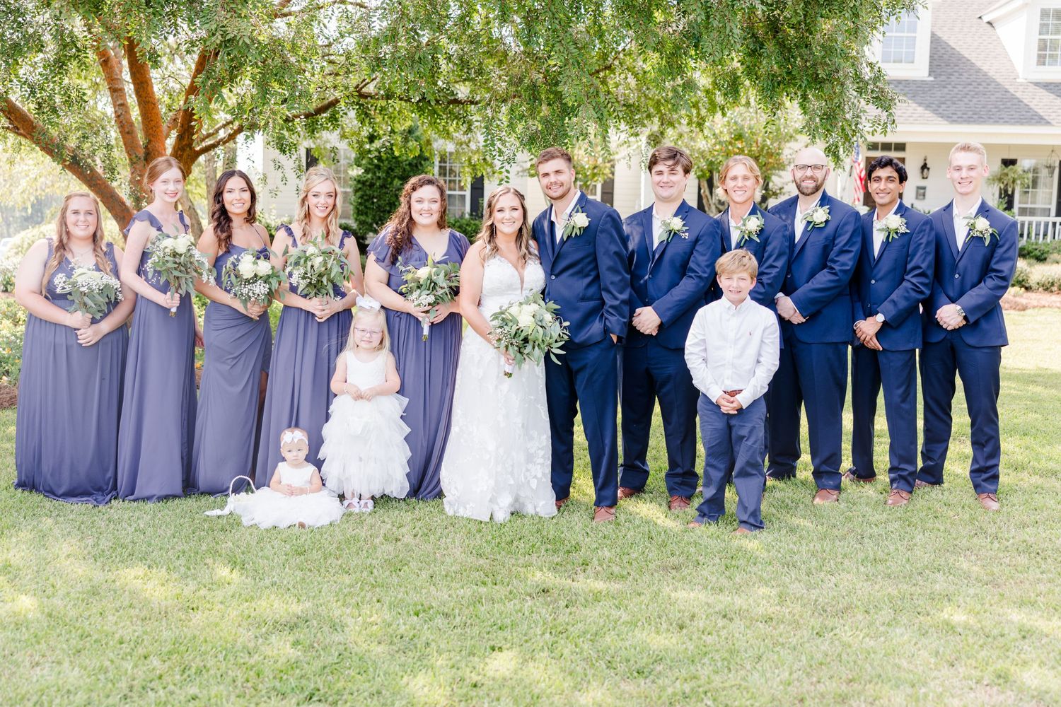 Wedding party in navy and lavender attire poses together on a sunny lawn.