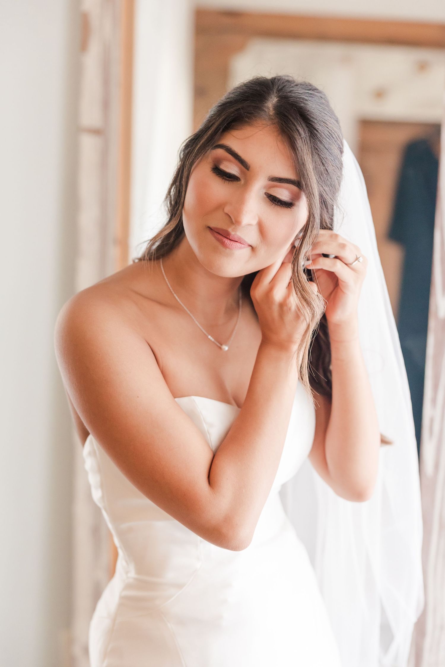 A bride in a white strapless wedding dress adjusts her veil and jewelry in an elegant indoor setting.