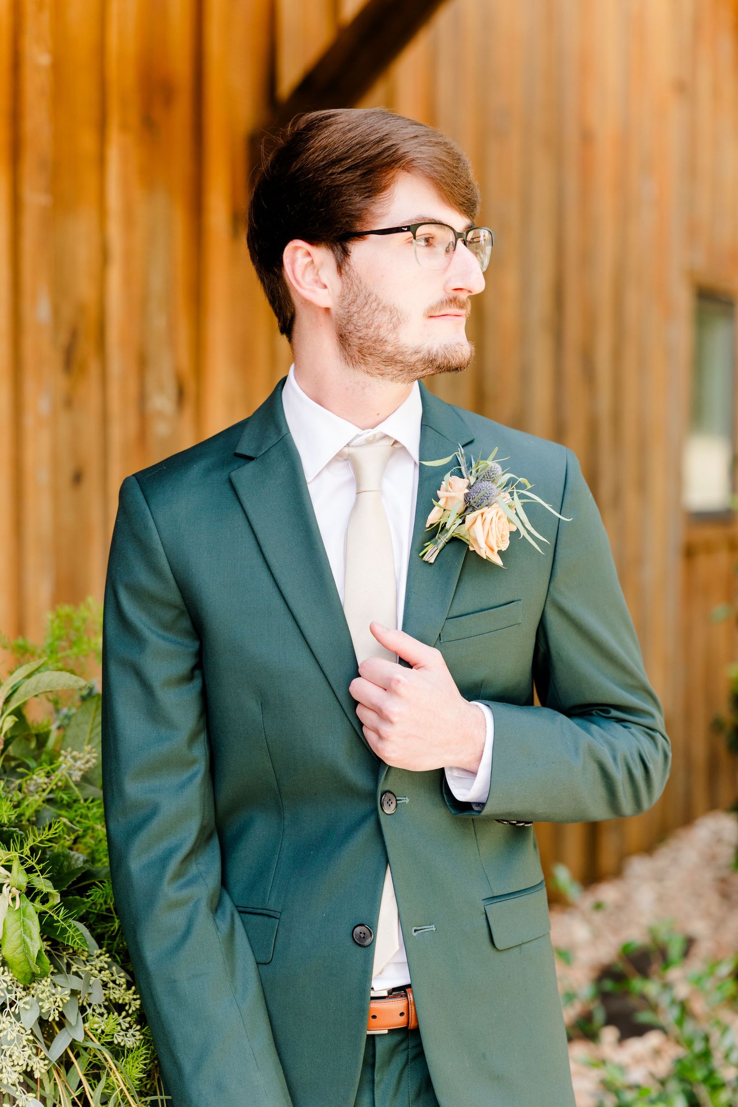 Groom in green suit with boutonniere and cream tie poses outdoors near wooden barn wall.