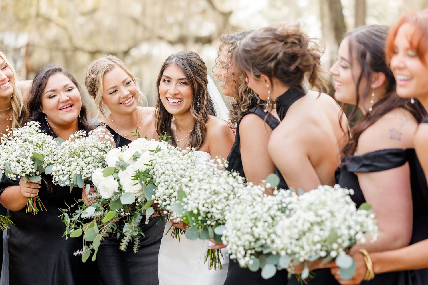 Wedding party members smile and laugh while holding delicate white bouquets.