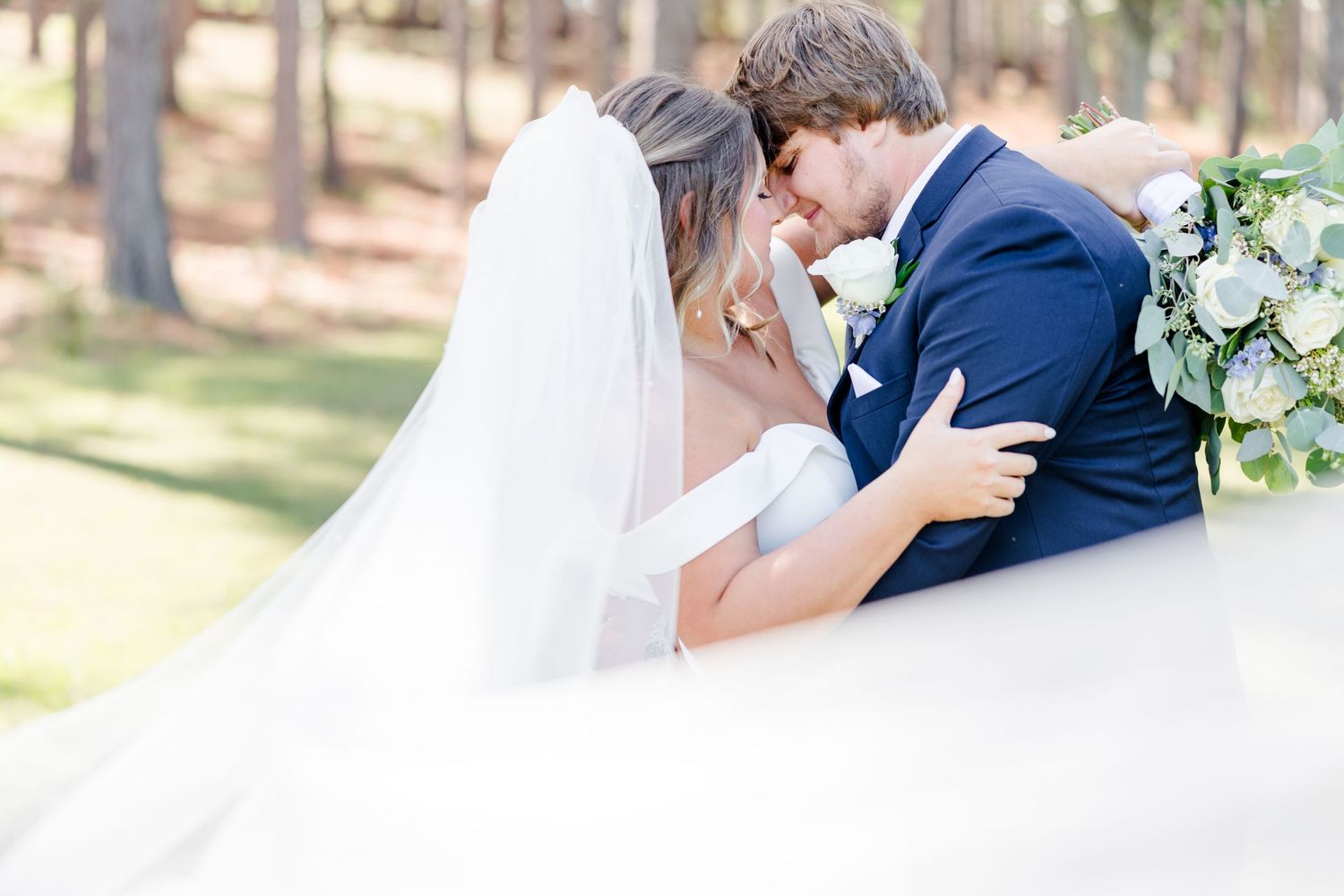 Dreamy wedding photo of couple embracing with flowing veil and forest backdrop.