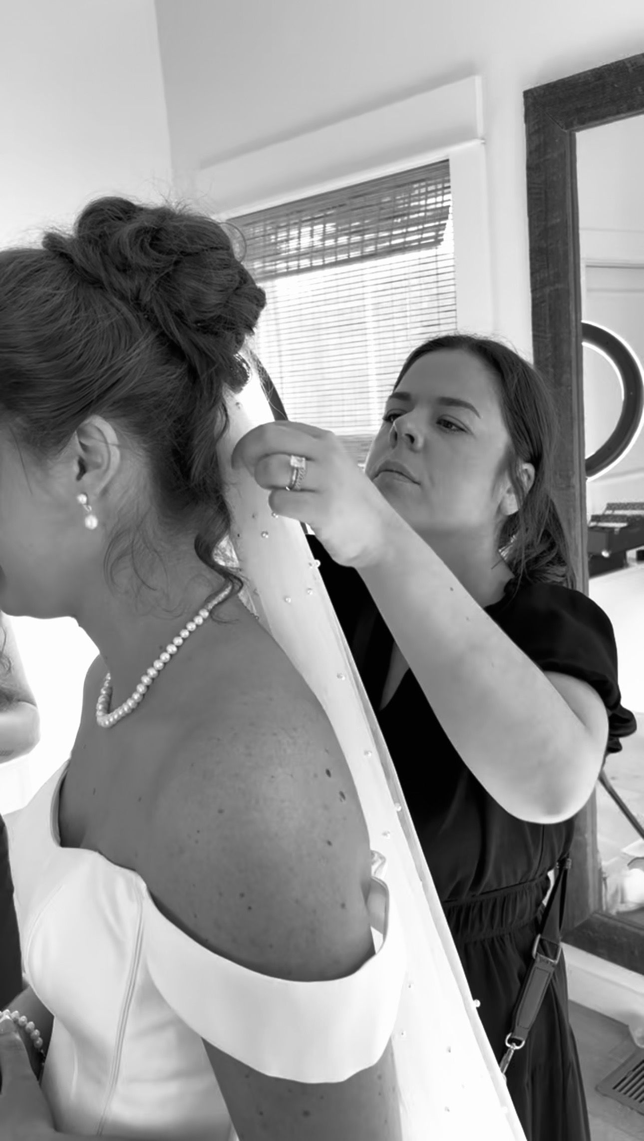 Black and white photo of a hairstylist pinning up a bride's elegant updo hairstyle before her wedding ceremony.