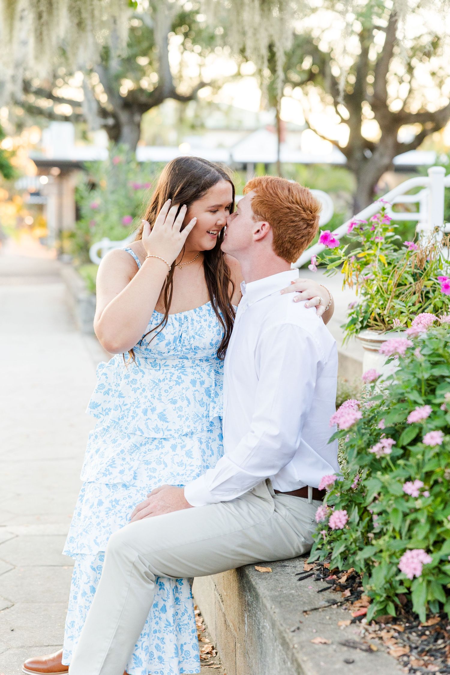 Couple sharing a romantic kiss outdoors, woman in blue floral dress showing engagement ring, surrounded by pink flowers.