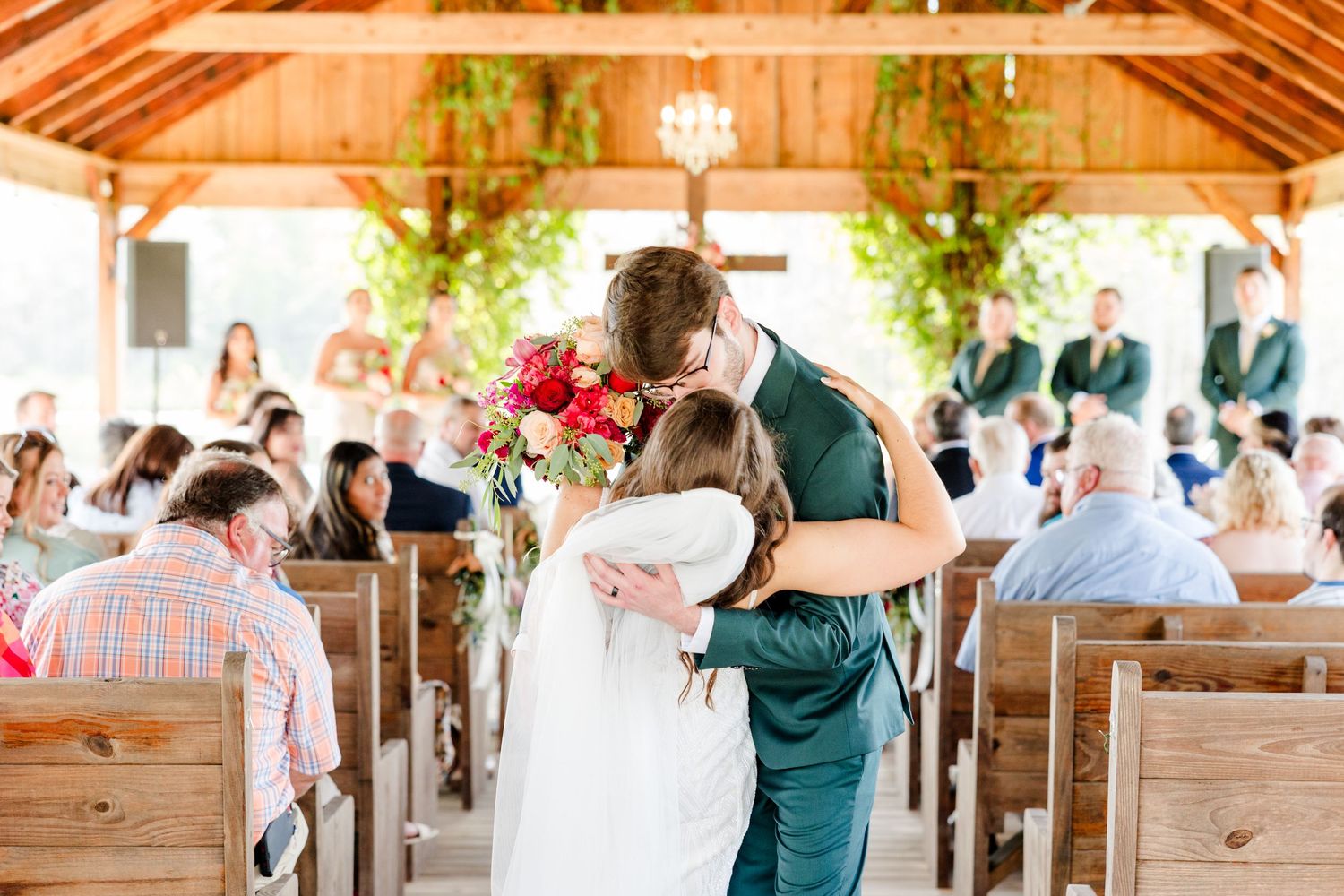 Bride and groom share first kiss at rustic outdoor wedding chapel with colorful bridal bouquet and seated guests.