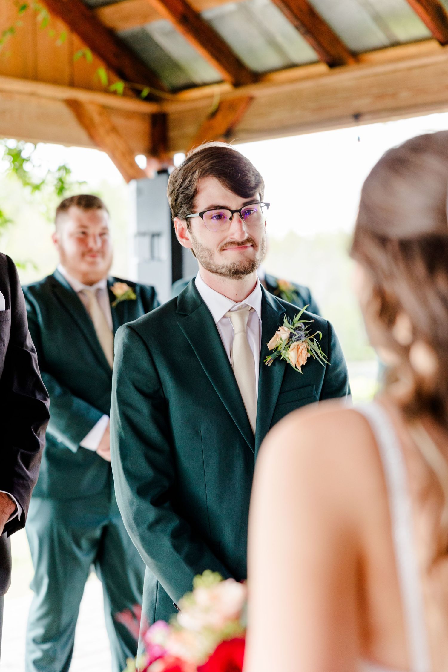 Groom in dark green suit smiling at bride during outdoor wedding ceremony under wooden pavilion.