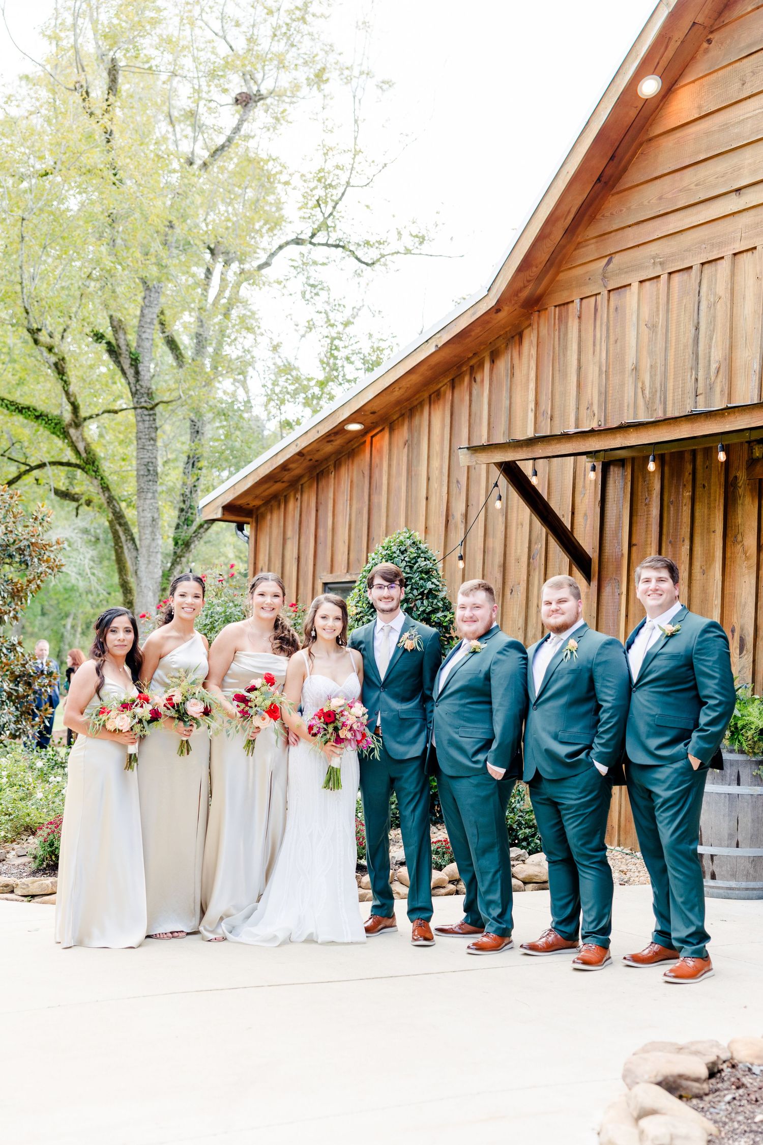 Wedding party posing outside rustic barn venue, bridesmaids in champagne gowns, groomsmen in teal suits holding colorful bouquets.