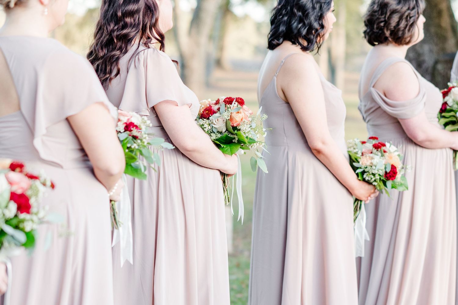 Bridesmaids in blush gowns holding colorful bouquets stand in a row outdoors.