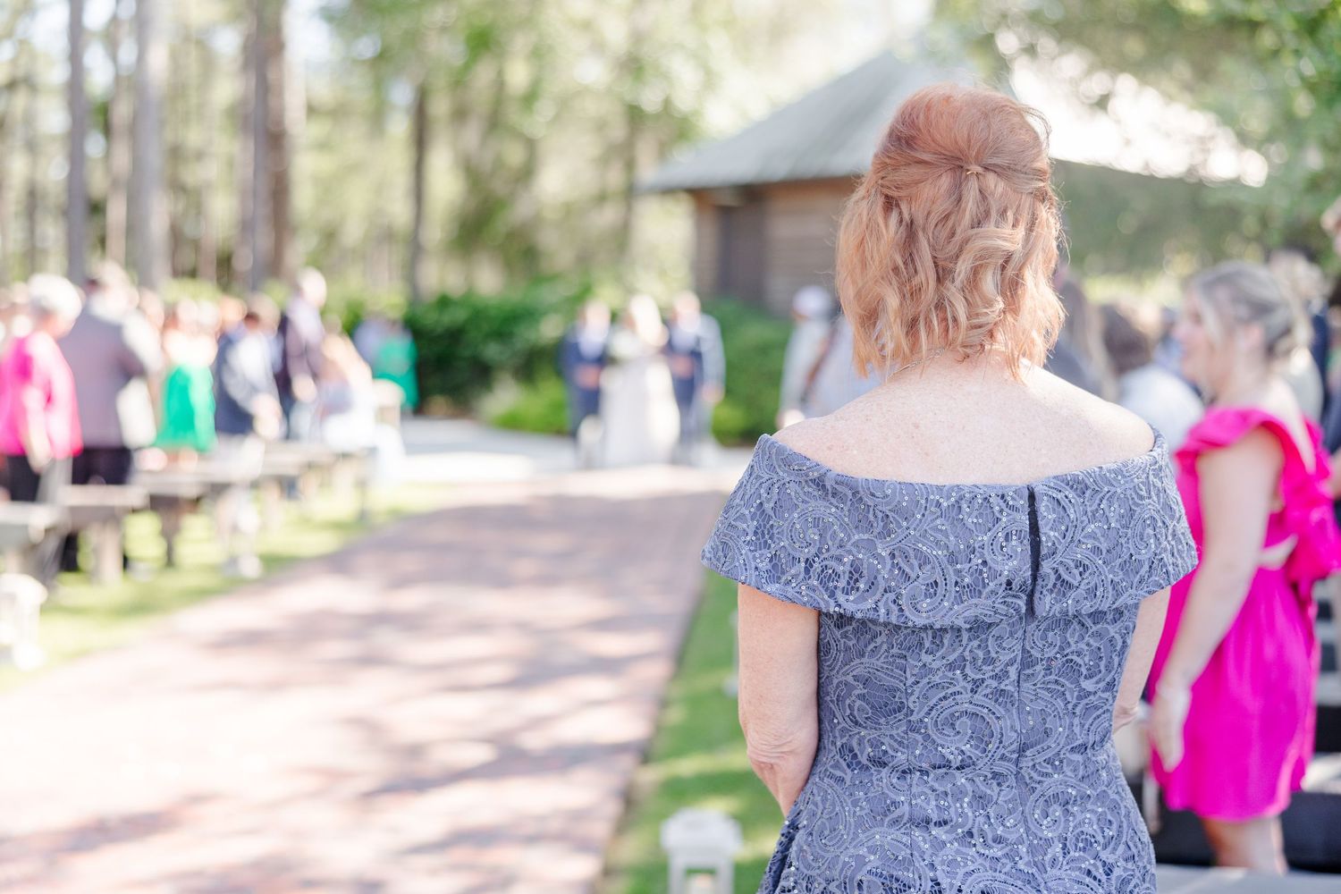 Wedding guests in bright dresses gather along a wooded outdoor ceremony path.