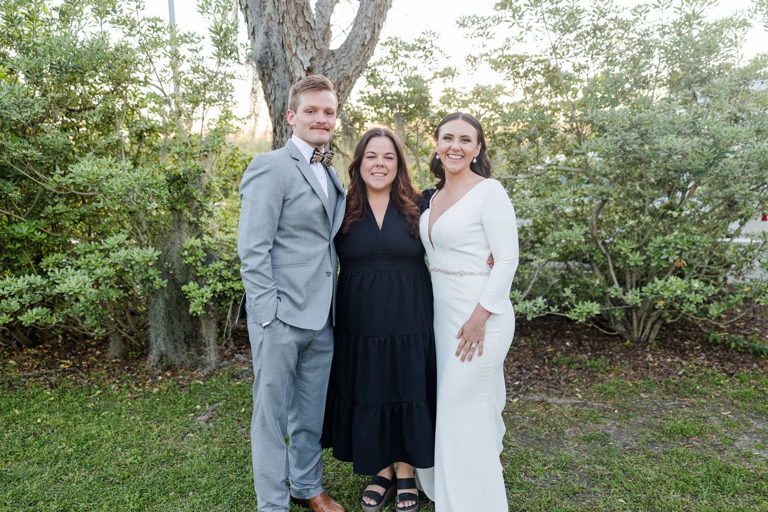 Three people in formal attire pose together outdoors against a green garden backdrop.
