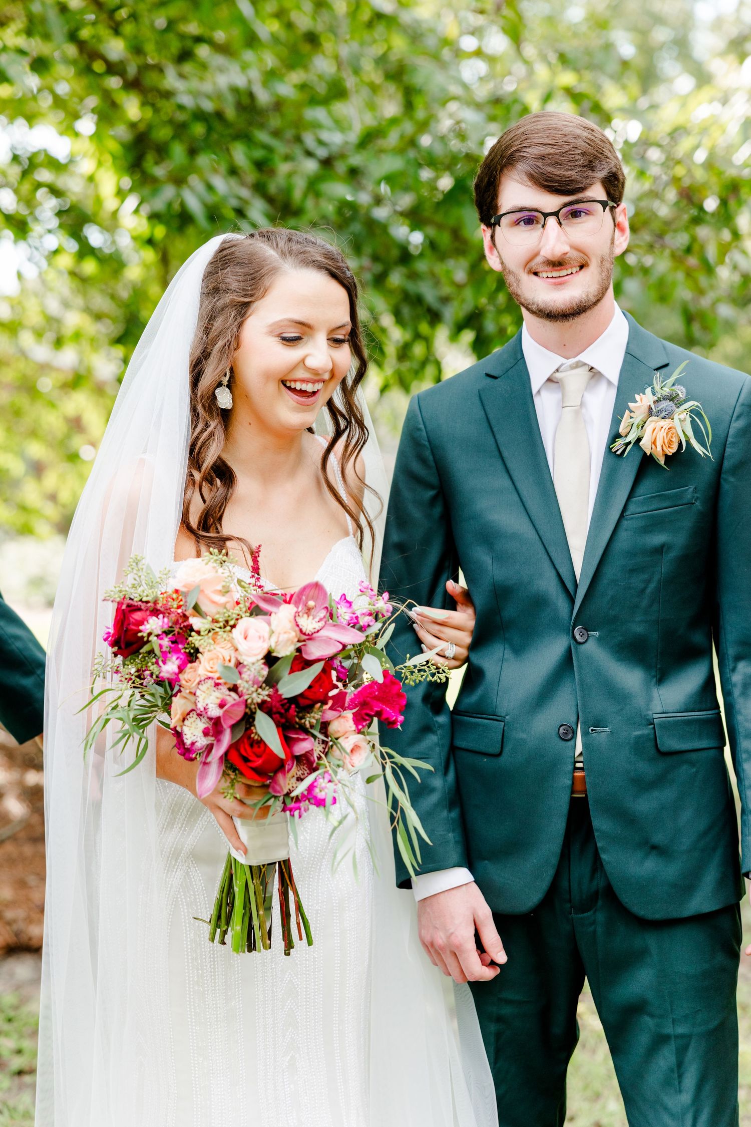 Bride holding vibrant pink floral bouquet stands smiling beside groom in teal suit with boutonniere outdoors.