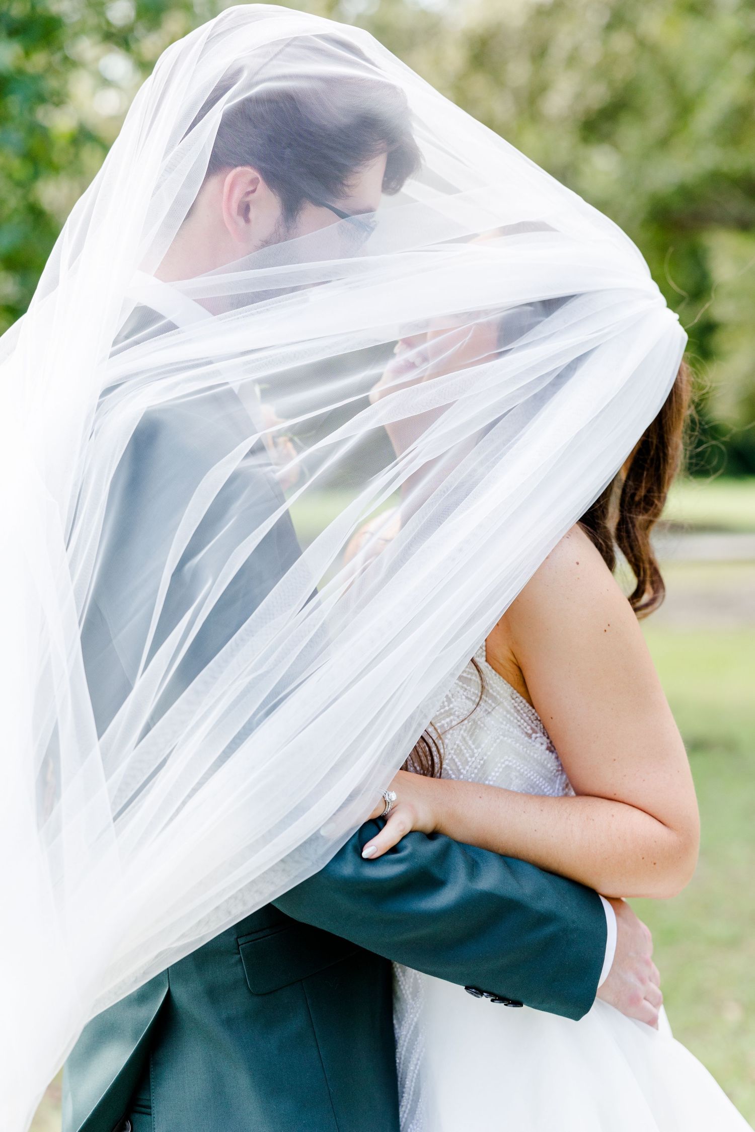 Bride and groom sharing a romantic kiss under a flowing white veil outdoors on their wedding day.