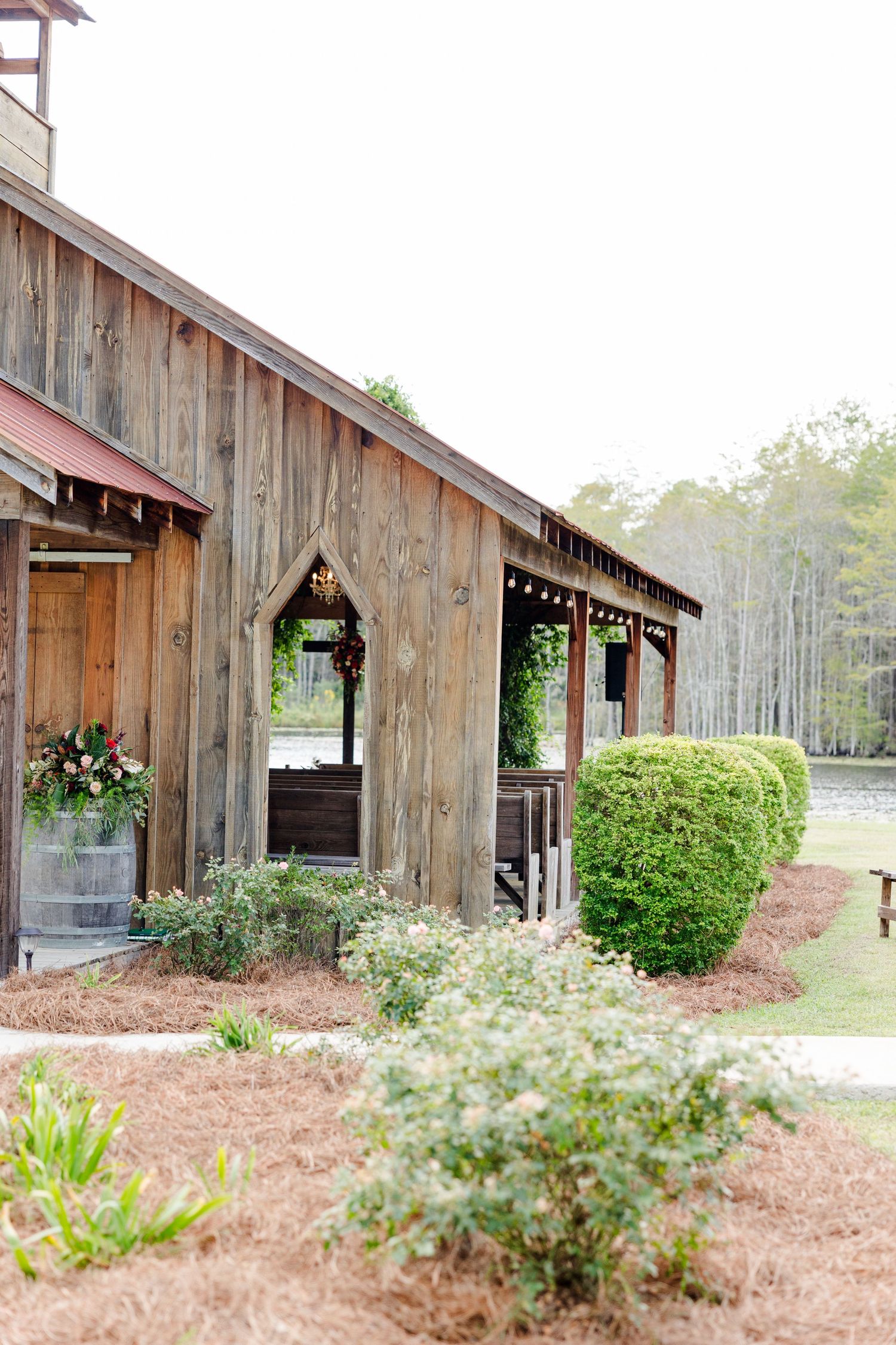Rustic wooden barn wedding venue with manicured hedges, pine straw landscaping, and floral decorations in a serene forest setting.