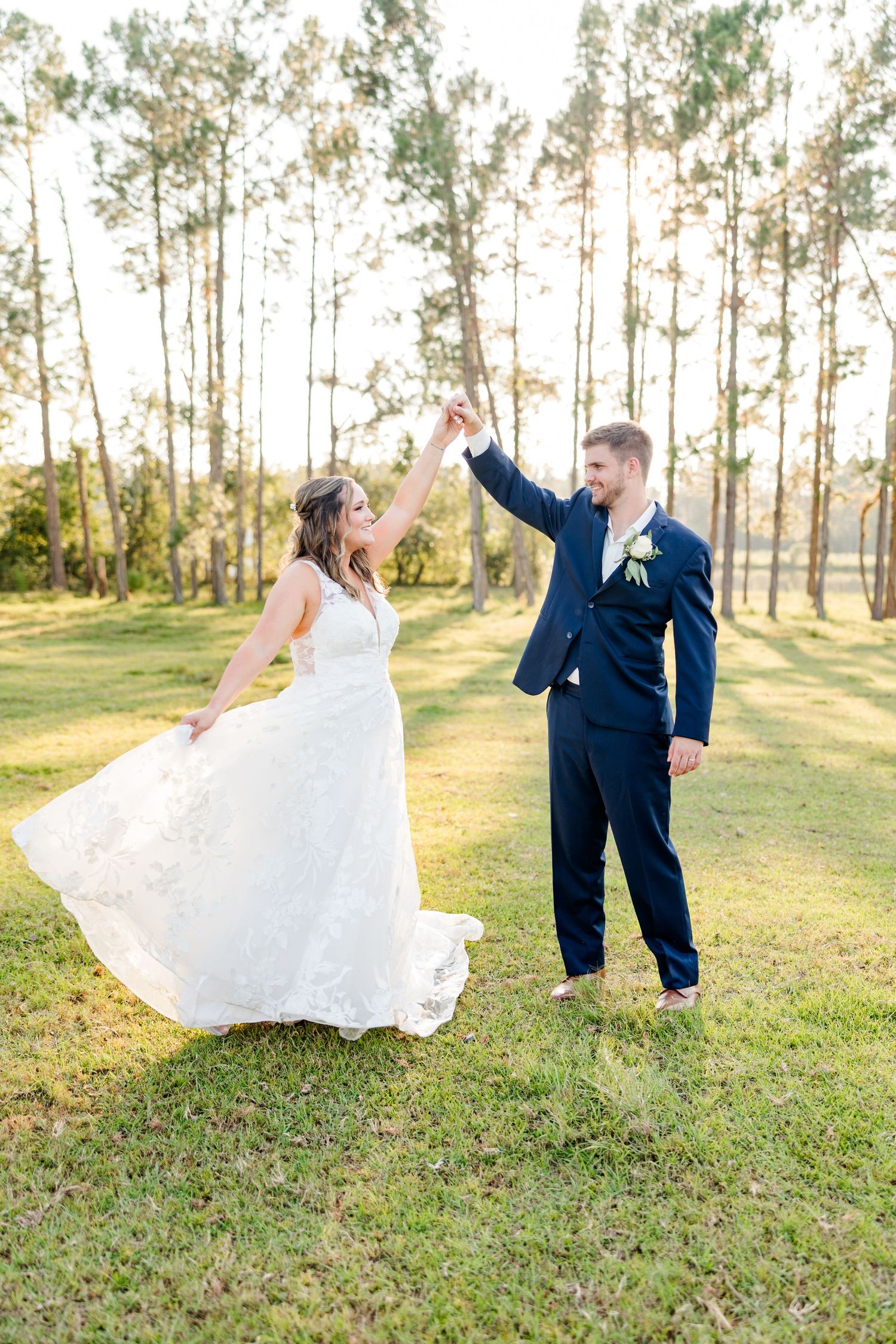 A couple dancing outdoors in wedding attire with tall pine trees in the background during golden hour.