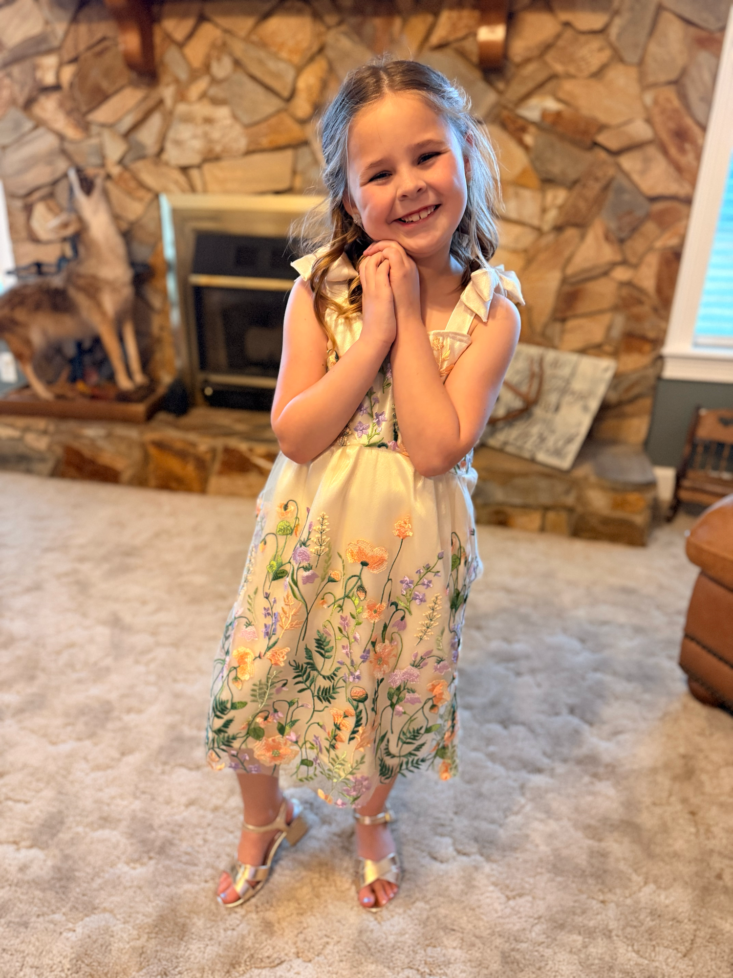 Young girl in floral embroidered dress with gold sandals smiling in front of stone fireplace.