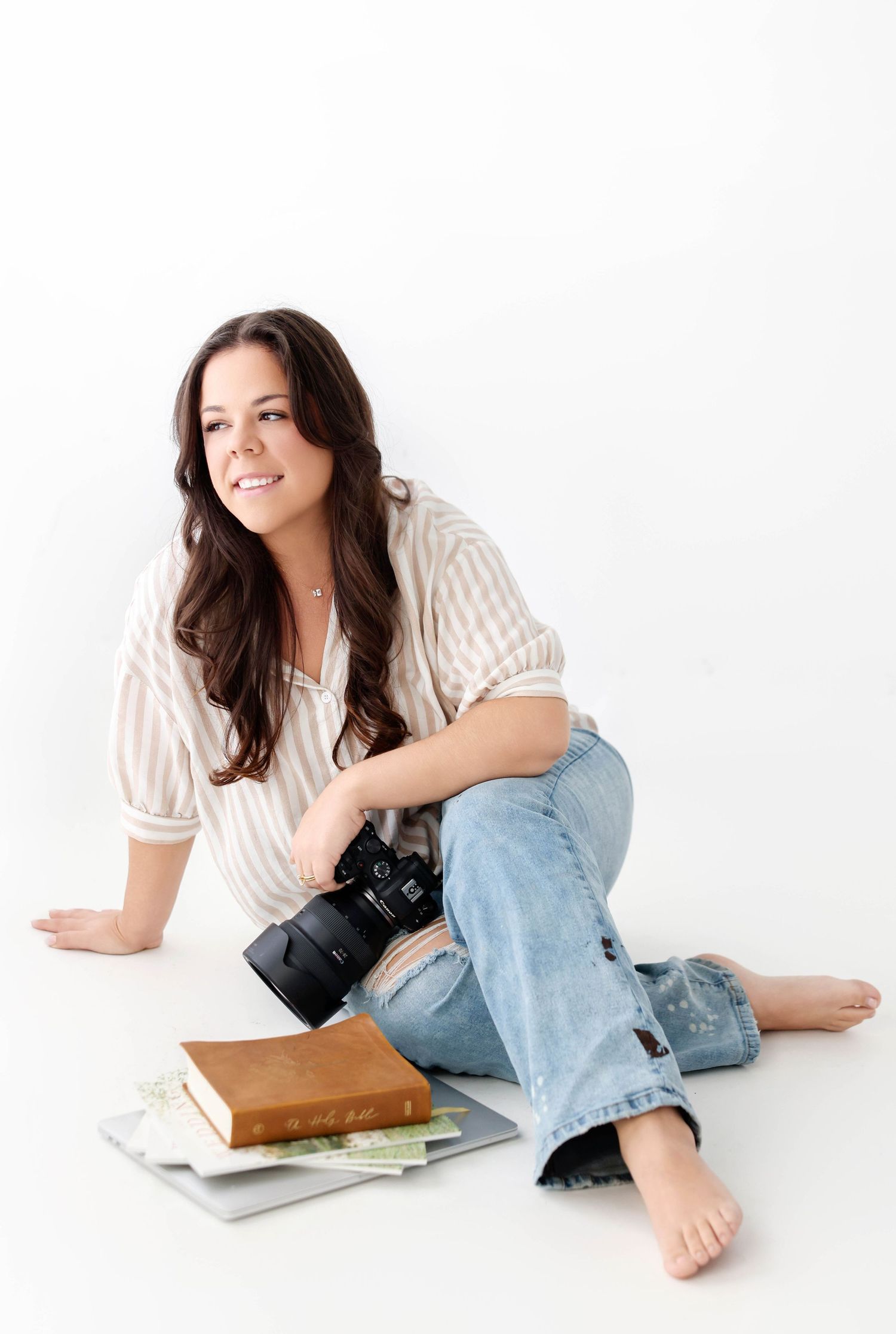 Photographer in striped shirt and jeans relaxes with camera and laptop on white floor.