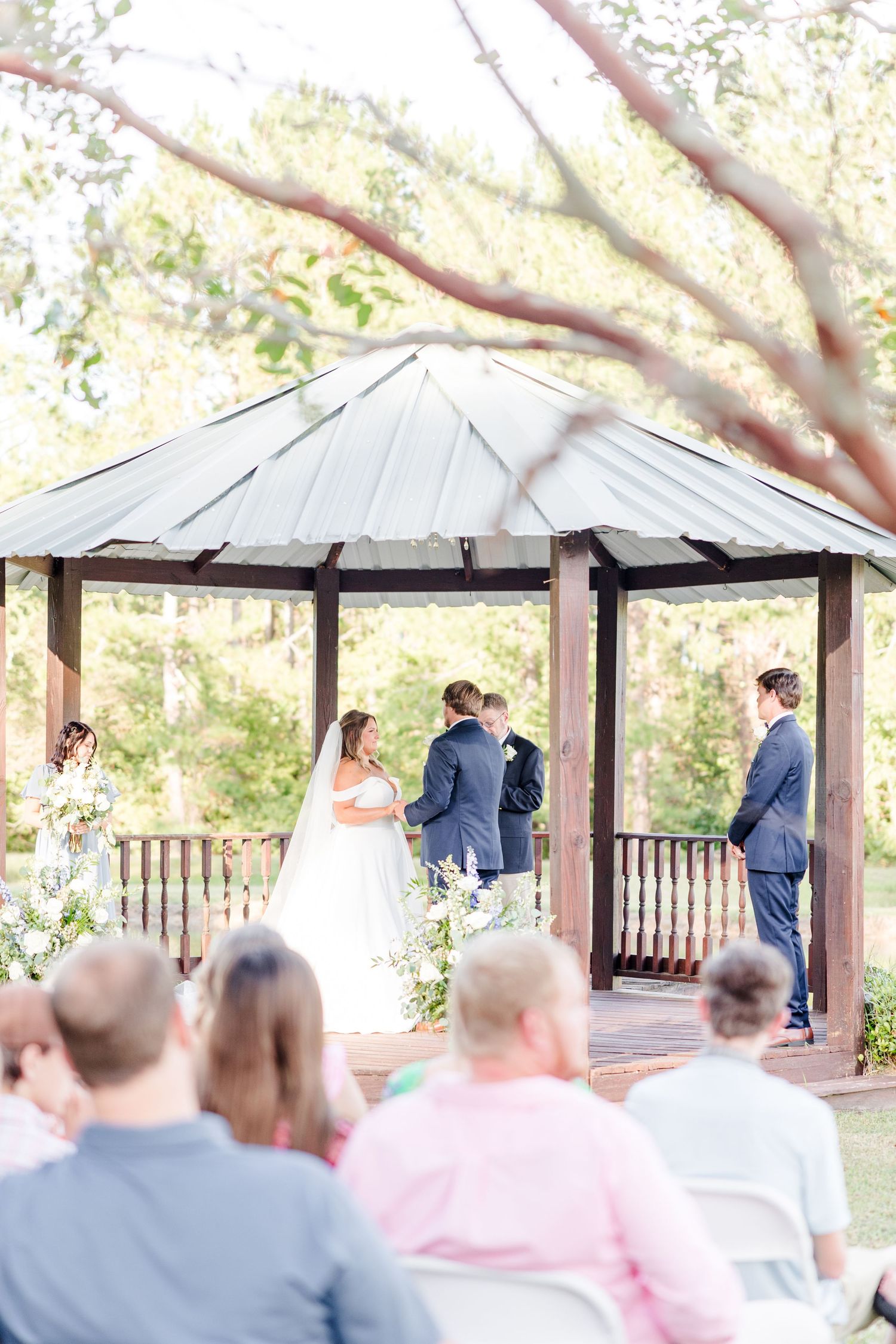 Wedding ceremony taking place under a rustic gazebo with wooden railings surrounded by trees.