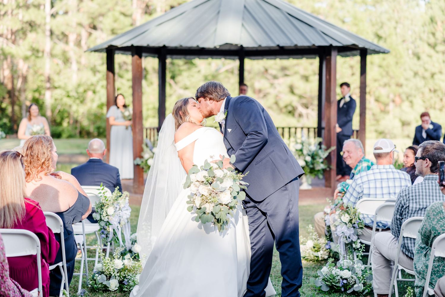 Wedding ceremony scene with guests seated outdoors under a wooden gazebo surrounded by greenery.