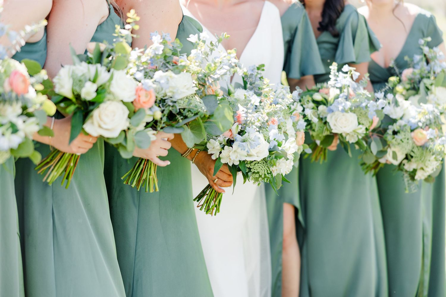 Row of bridesmaids in sage green dresses holding white and green floral bouquets.