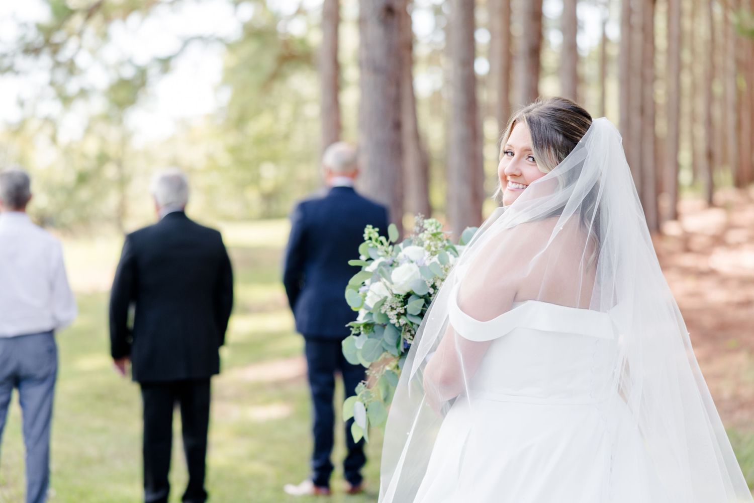 Outdoor wedding ceremony setup among tall pine trees with guests gathering.