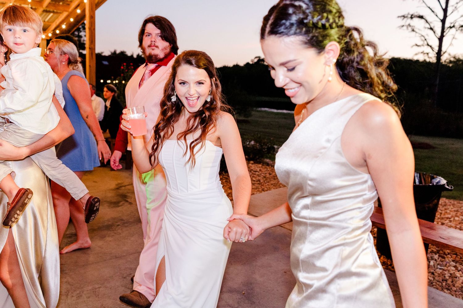Two brides in white gowns joyfully dancing together at an outdoor wedding reception at dusk.