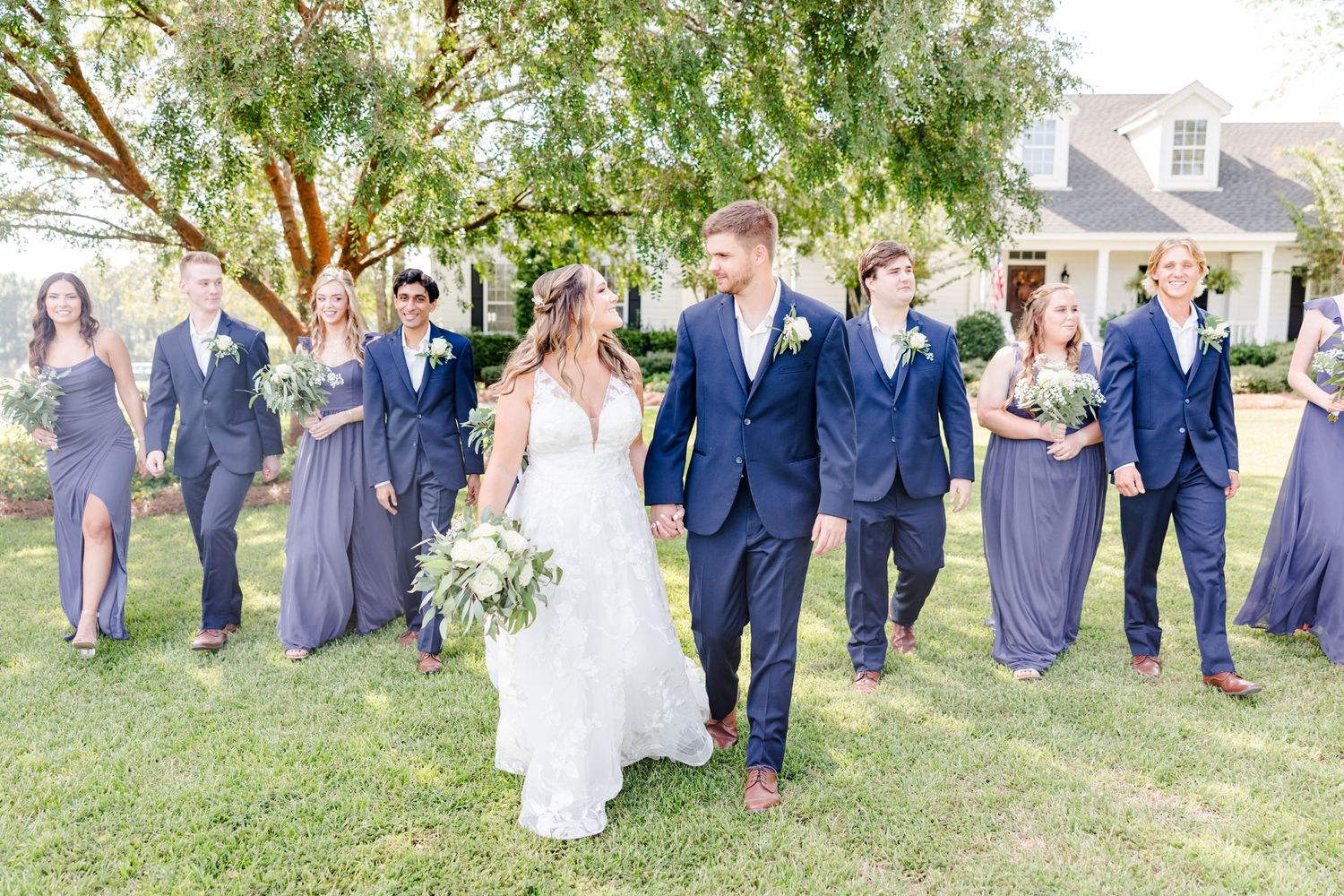 Large wedding party in navy and purple poses for group photos on grass.
