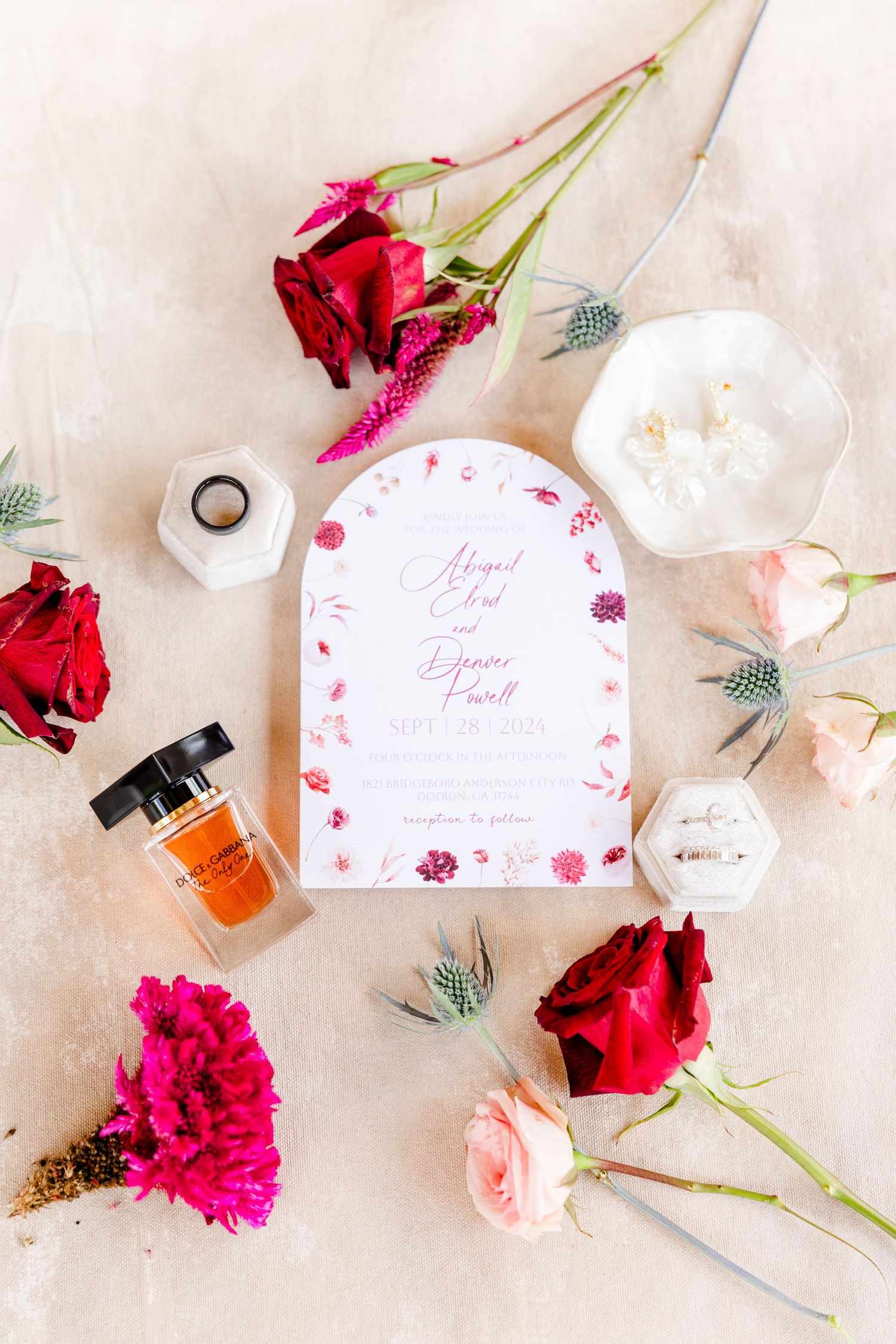 Elegant wedding invitation suite with red roses, carnations, perfume bottle, and ring box on a neutral background.