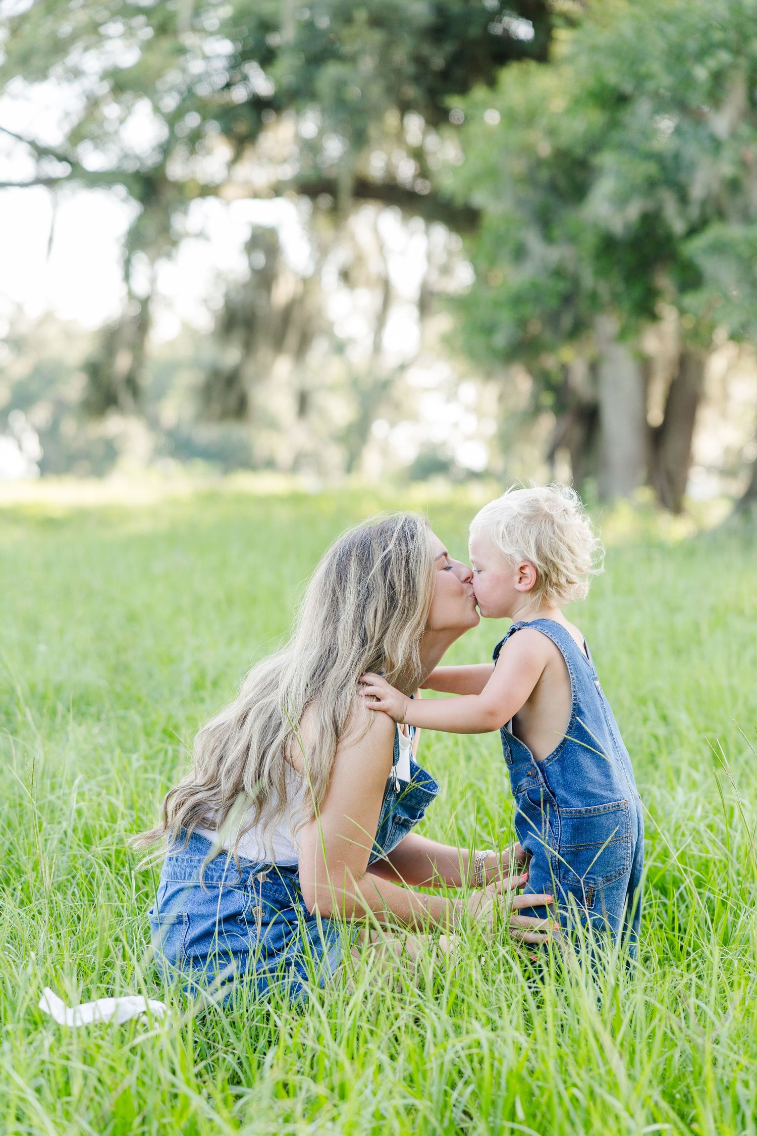 A mother and toddler share a sweet kiss sitting in lush green grass, both wearing denim overalls outdoors.