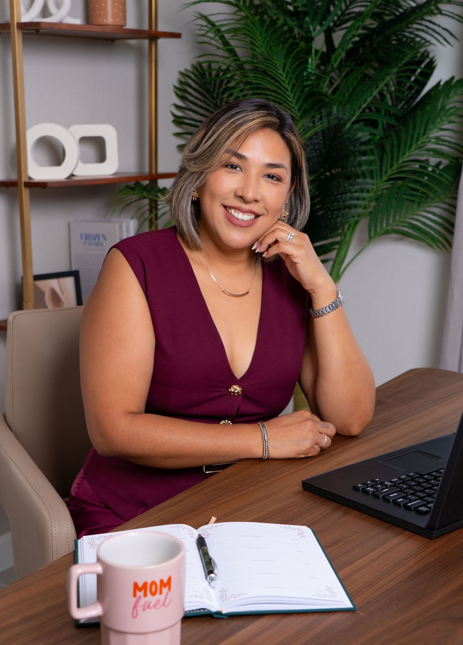 Professional woman in burgundy dress smiling at desk with laptop, notebook, and Mom Fuel mug in modern office.