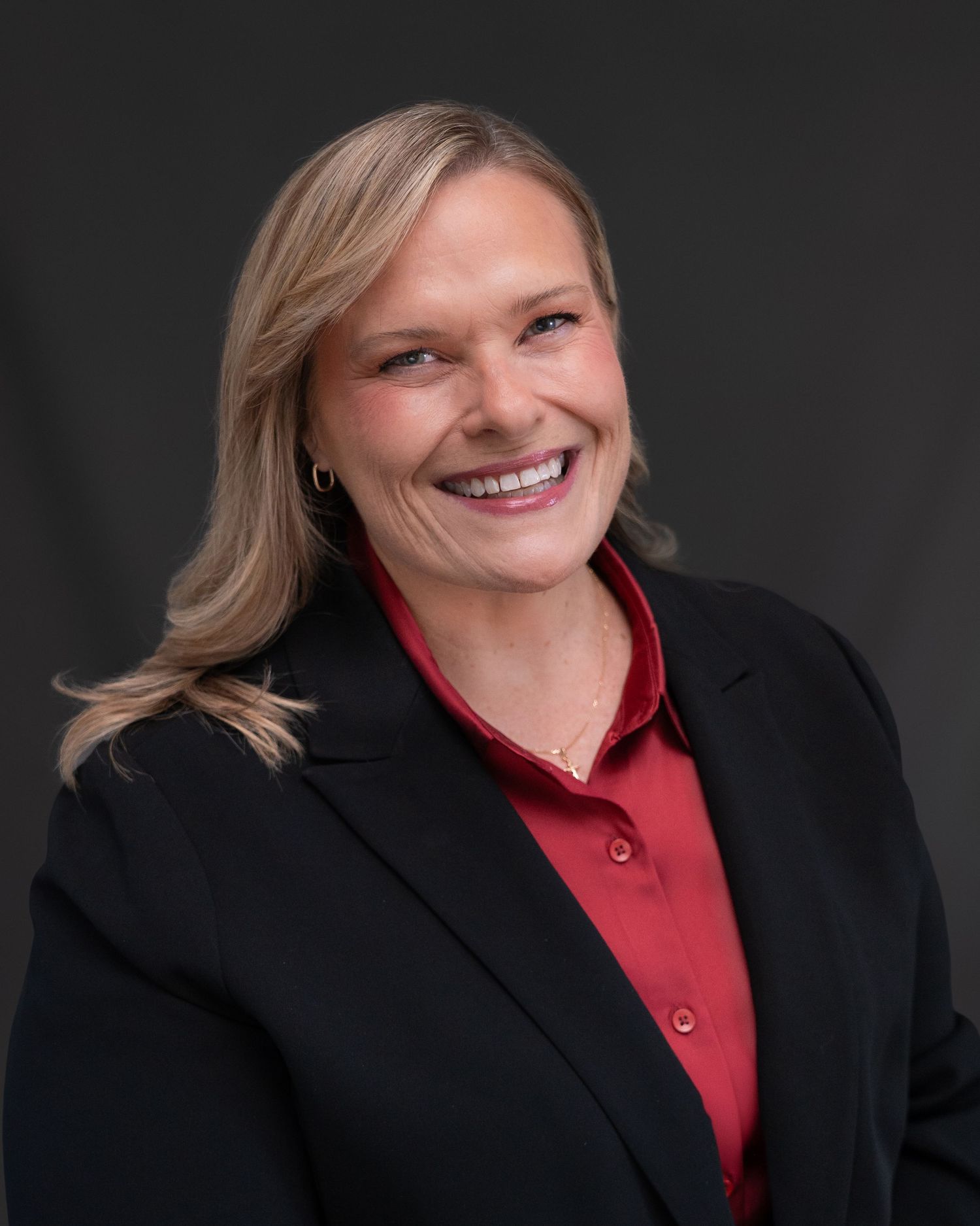 A smiling blonde woman in a black blazer and red shirt poses for a professional headshot.