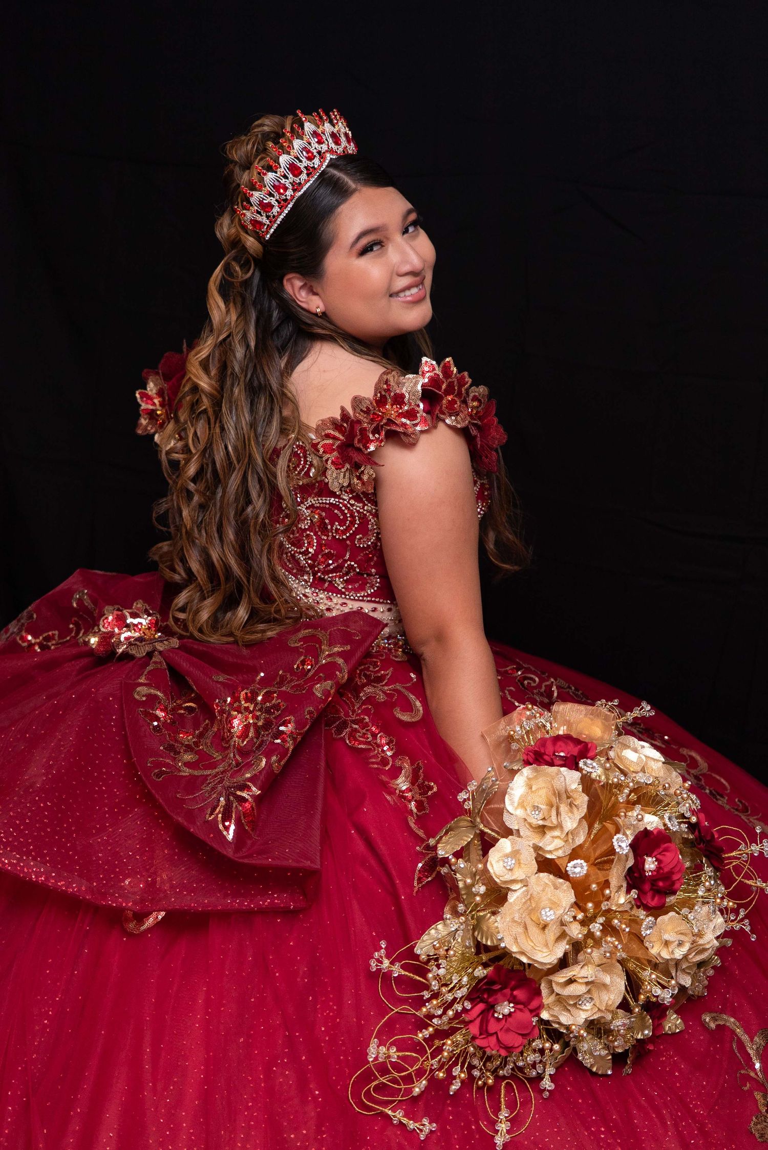 Teen girl in red and gold embroidered quinceañera gown with ruby tiara holding gold roses bouquet.