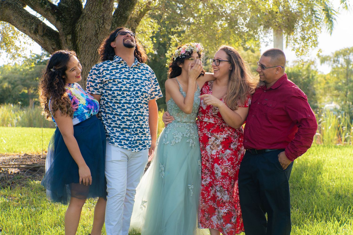 Joyful family of five laughing together outdoors under a tree during a sunny day portrait session.