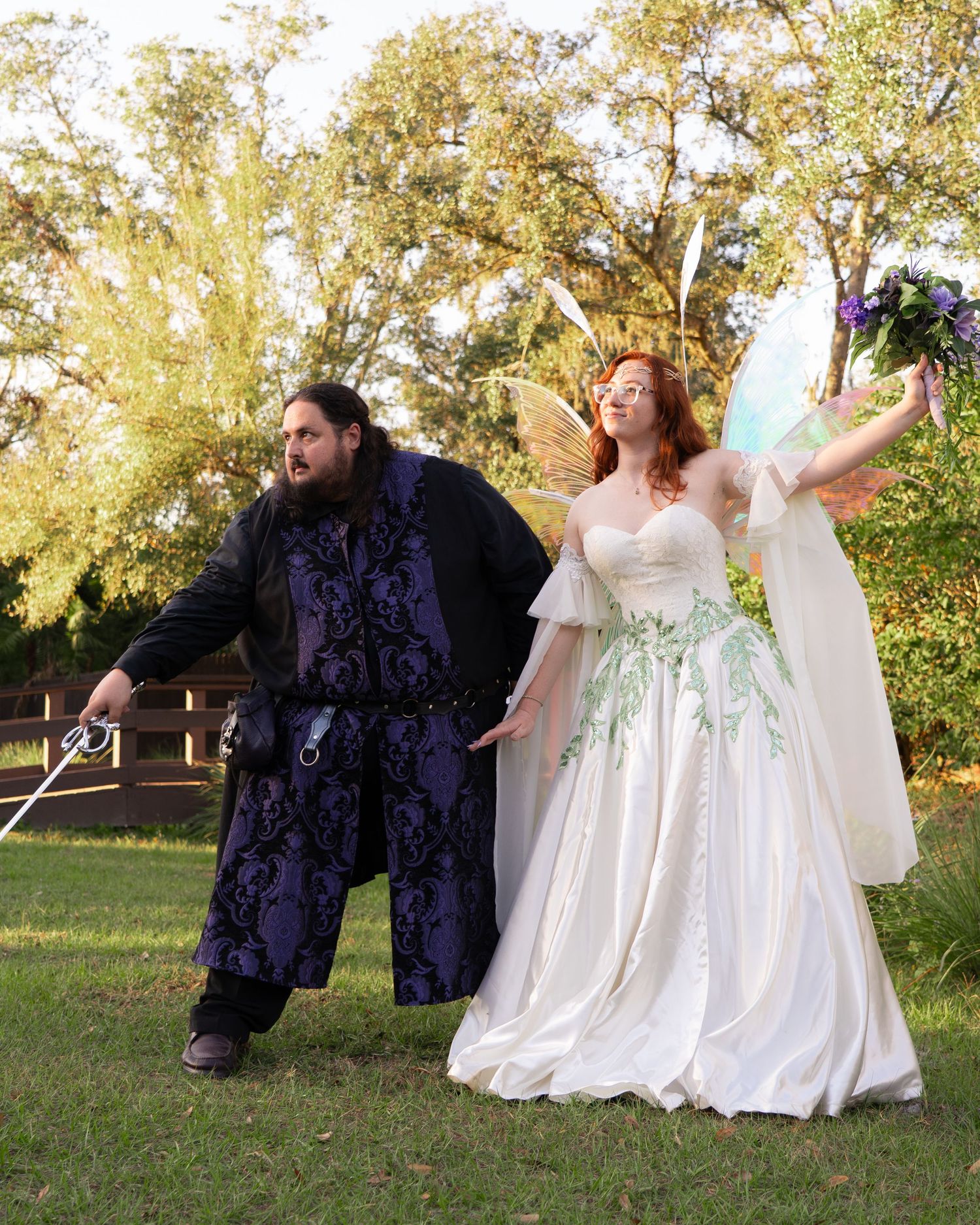 Couple in fantasy wedding costumes outdoors, bride in fairy wings and floral gown, groom in dark medieval attire.