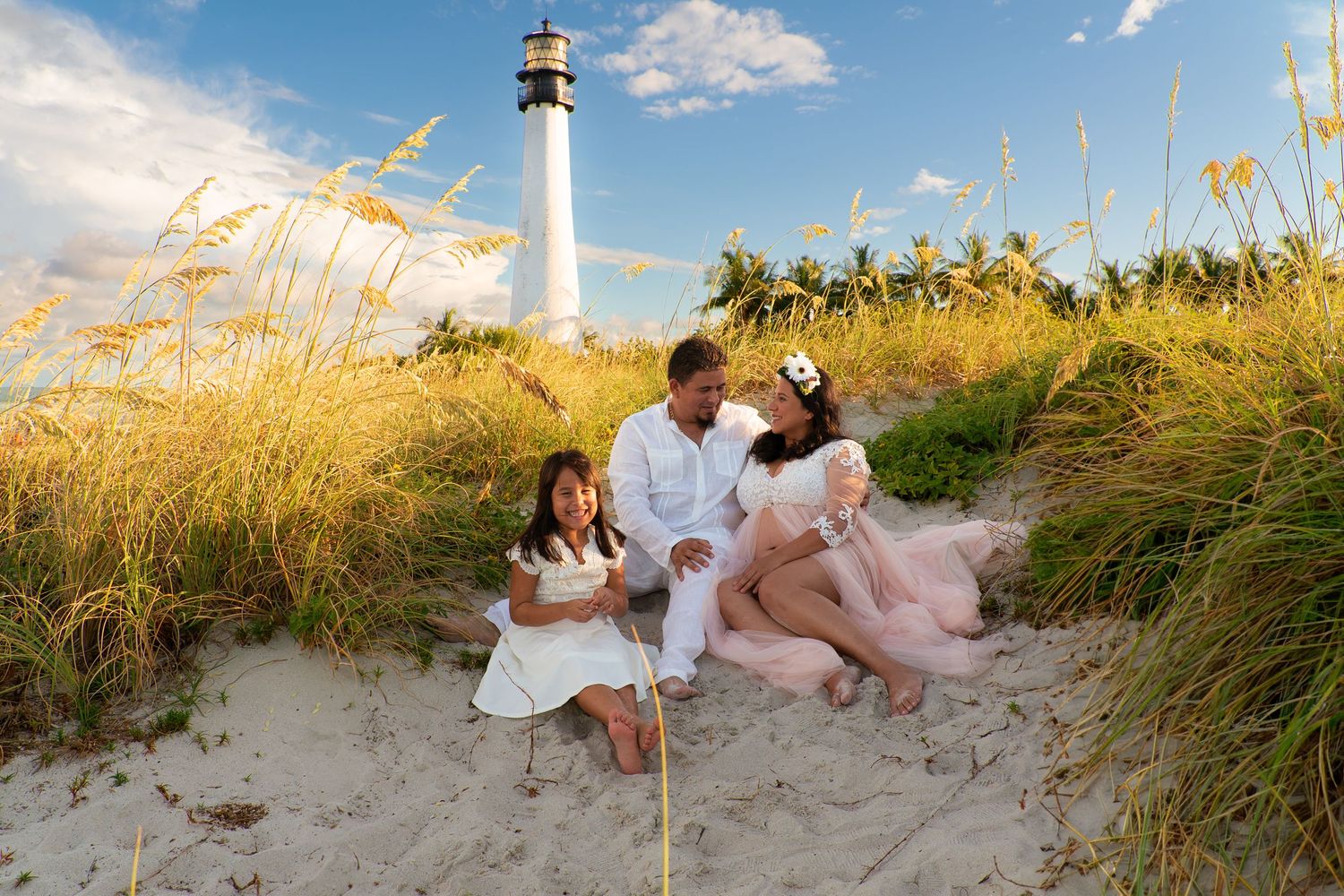 Family sitting on sandy beach dunes near a white lighthouse at sunset.