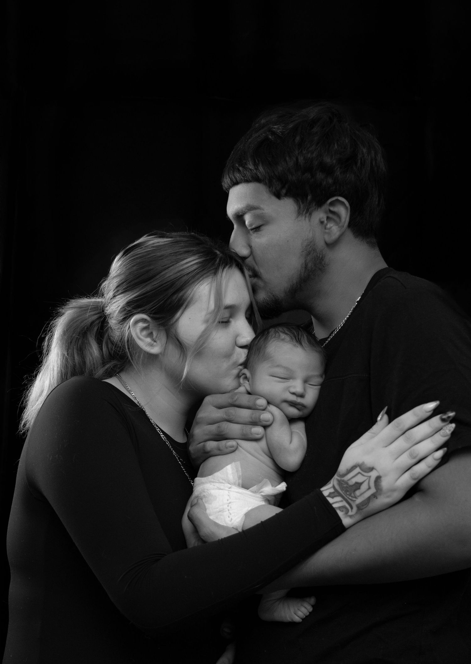 A black and white portrait of two parents tenderly kissing their sleeping newborn baby.